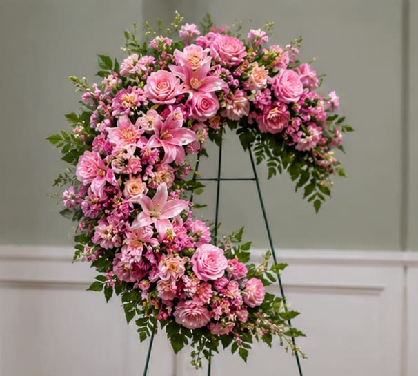 Heartfelt Love Pink Sympathy Wreath - A soft, romantic standing sympathy arrangement-this Heartfelt Love pink sympathy wreath makes a graceful memorial statement for funerals or celebration-of-life services. The curved, open-circle design and pale pink palette send a tender message of love and remembrance, ideal when you want the person you're thinking of to feel honored and comforted.  Densely packed with pink roses, pink lilies, carnations and smaller clustered blooms in blush and rose tones, the wreath is woven with green foliage for contrast and texture. Mounted on a metal easel stand and shaped as a large floral ring, its formal scale suits casket-side displays or chapel settings rather than a handheld bouquet.  Choose this wreath to express sympathy, deep affection, or to mark an anniversary of a loss-the soft colors and lush flowers convey warmth and consolation. The arrangement arrives ready to display on the included metal easel; lilies and roses are best kept cool and away from direct heat to extend vase life during services.