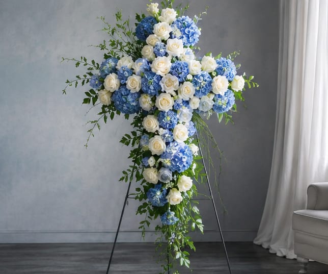 Heavenly Sky Funeral Cross  - A serene blue-and-white standing spray shaped like a cross, designed for sympathy and memorial services to honor a life with quiet dignity. The Heavenly Sky Funeral Cross uses clusters of powder-blue hydrangeas and pure white roses to create a calm, ceremonial silhouette that reads both reverent and comforting to the person you're remembering.  Densely packed blooms form the upper crossbar and central focal area, tapering into a trailing vertical section framed by airy green foliage for a gentle, flowing profile. The cool blue and crisp white palette conveys peace and respect; roses add classic formality while hydrangeas bring soft volume and texture. Mounted on a sturdy easel, the arrangement stands tall for viewing near a casket, memorial table, or service altar.  A meaningful way to express sympathy and support for grieving family and friends, this cross-shaped spray helps convey love, remembrance, and solace at a funeral or memorial. Sized for prominent display on the included easel, it arrives ready to place with simple handling; no additional care is required beyond routine water refresh if kept before the service.
