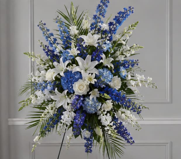 Sea Side Remembrance Funeral Spray  - Blue and white blossoms arranged with coastal calm create a dignified sympathy spray that reads as both peaceful and reverent-the perfect floral tribute for a funeral or memorial service. This funeral standing spray is designed to convey comfort and respect, so the person you're honoring will be remembered with a serene, graceful presence at their service.  The arrangement pairs large clusters of blue hydrangea with white roses, white lilies, and delicate white daisies, while tall blue delphinium-like stems add vertical movement and an airy, lifted feel. Textural contrast comes from spiky white blooms and elegant orchids tucked among the blooms, with long palm fronds and lush greenery fanning outward for a full, formal silhouette. A tasteful blue ribbon and a sturdy floral stand on a standing easel complete the ceremonial presentation.  Appropriate for a church service, graveside memorial, or any sympathy setting, this spray reads as a calming statement of remembrance for family members and friends. It arrives ready to display on its easel; for best longevity, keep the arrangement in a cool location out of direct sun until the service.