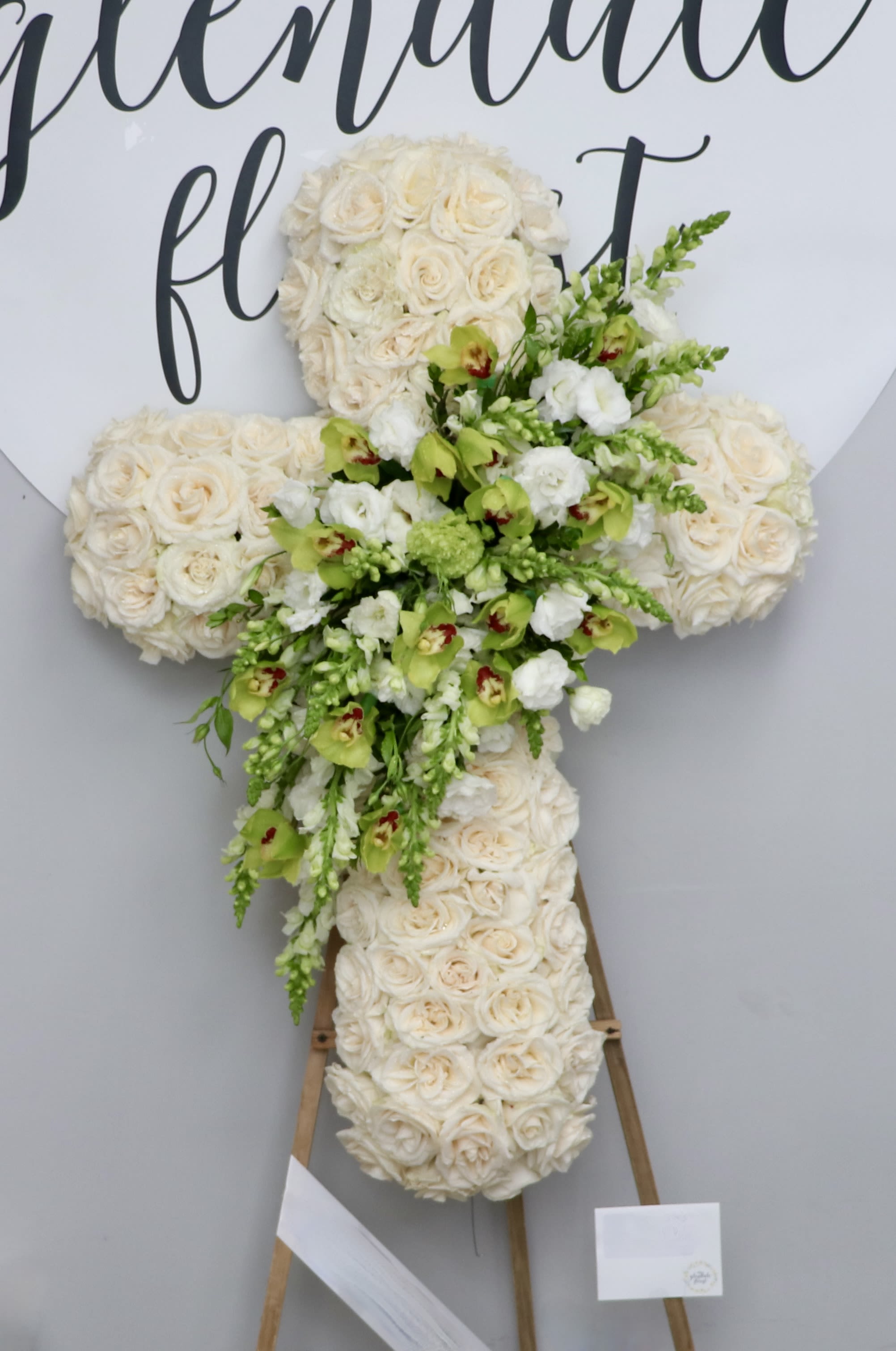 White Cross and Orchids - This cross is made with white roses and includes a design of green orchids and other seasonal greens.    We include easel, printed banner, and delivery (some fees may apply). An additional banner can be added for $15.    Standard size is 30'', deluxe is 36'', and premium is 42''.