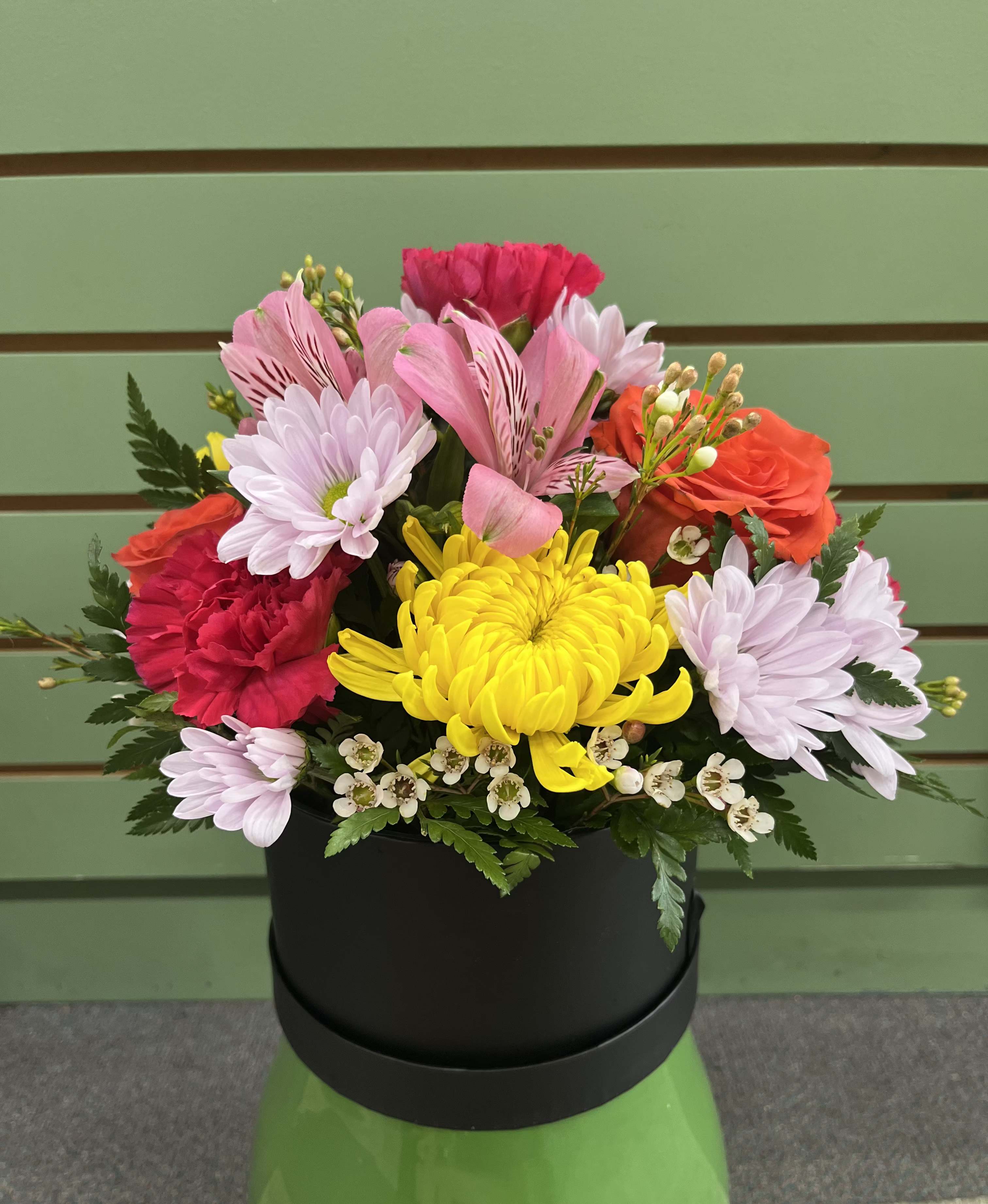 Brightest Blooms Hat Box - This arrangement combines cheerful yellow chrysanthemums with pink alstroemeria, soft lavender daisies, hot pink carnations,  and orange roses. Designed in a modern black hatbox, the contrast makes the colors pop and suits spring or summer celebrations as easily as holiday gifting.  Pretty Petals brings a burst of joyful color for a birthday, just because, or thinking-of-you moment. The rounded composition reads as bright and celebratory, so the person you're sending it to will feel noticed and genuinely cheered whether you're marking a milestone, saying thank you, or brightening a room after an illness. It is also perfect for anniversaries, housewarmings, retirements, ideal for a friend, parent, partner, or colleague. The compact design fits tabletops and entryway consoles.