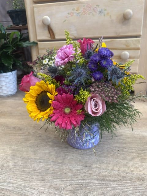 Summer Symphony  -  A lively explosion of peak-season warmth that perfectly captures the carefree joy of a sun-drenched July afternoon. Bring the radiant energy of the outdoors inside.  Arrangement will either come in a pink or purple container as shown. 