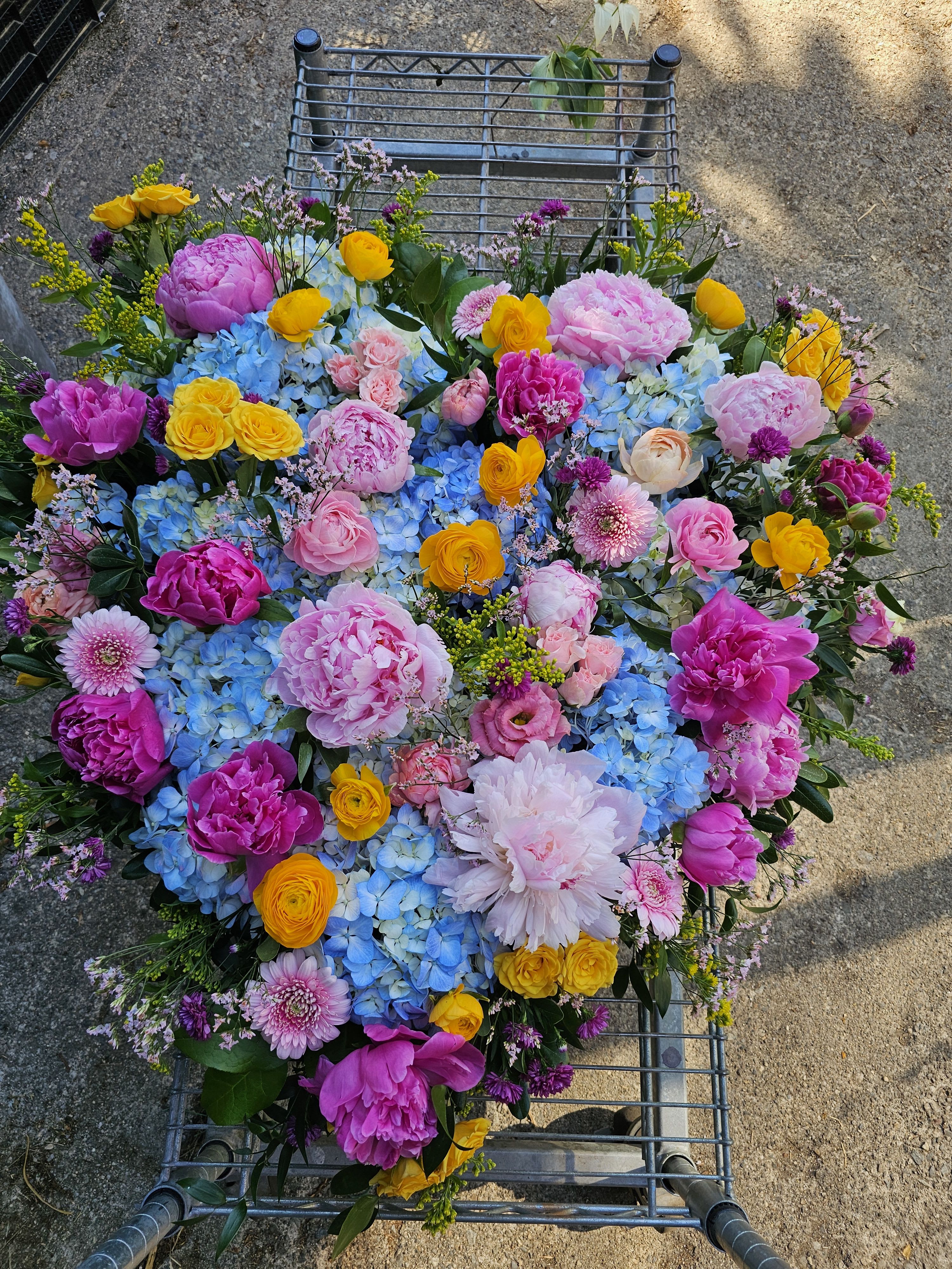 Heart of a Lifetime Tribute  - A lush, heart-shaped tribute overflowing with large peonies and ranunculus makes an immediate, romantic impression-perfect as a sympathy tribute or a statement piece for a memorial service. This flower arrangement heart combines pinks, cheerful yellows, soft blues and touches of lavender to create a warm, celebratory tone that honors a life while offering comfort to those who receive it.  Densely packed with layers of texture, the design pairs bold blue hydrangea clusters with small spray roses, daisy-like accents and other seasonal blooms to form a deeply dimensional surface. The colors read as both gentle and uplifting, and the mix of full peony heads and delicate ranunculus blossoms gives the piece a tactile, garden-fresh feel. The finished heart is presented on an easle highlighting its large scale and making it easy to display at a service or event.  For the person you want to remember, this arrangement says love, warmth, and celebration of a life well-lived-an especially fitting choice for memorials, funeral services, or a tribute at a celebration of life. 