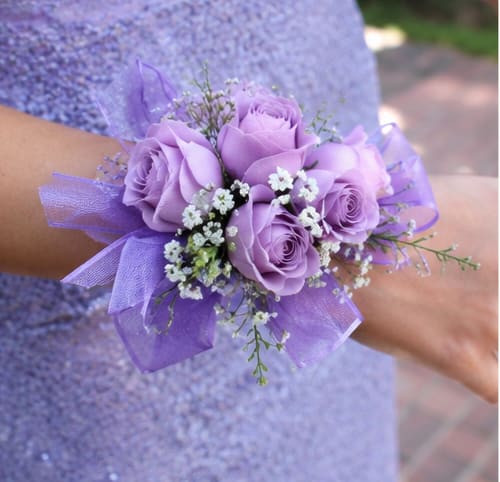 Simple Lavender Corsage  - Lavender roses clustered into a compact wrist corsage give an instant touch of romance-perfect as a prom corsage, bridal party accessory, or a thoughtful gift for Mother's Day or an anniversary. The Simple Lavender Corsage frames the wrist like a delicate bracelet, sending a soft, elegant message that the person you're thinking of is cherished and celebrated.  Tightly arranged lavender roses are accented with small white filler blooms and wisps of airy purple ribbon, finished on a secure wrist corsage band for comfortable wear. The color palette of purple, white, and hints of pink creates a refined, feminine look that pairs beautifully with formalwear or a springtime dress.  Made to be lightweight and polished for all-day wear at weddings, dances, or special events, this corsage arrives ready to fasten and enjoy. For best longevity, keep cool and mist lightly with water if needed; please request same-day delivery when ordering for last-minute occasions.