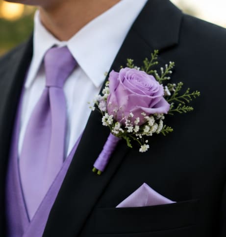 Simple Lavender Rose Boutonniere  - A single lavender rose boutonniere that reads as quietly romantic-perfect for a wedding boutonniere or other dressy celebration. The soft purple bloom is paired with delicate white filler flowers and sprigs of greenery to create an elegant, coordinated look that complements a black suit jacket and a matching lavender tie or pocket square, giving the person you're honoring a thoughtfully styled, romantic finish.  This compact arrangement is tailored for formal wear: one lavender rose with small white accents and green sprigs, secured with a boutonniere wrap for neat pinning to a suit lapel, dress shirt, or jacket. Sized to sit subtly on the lapel, it's ideal for grooms, groomsmen, fathers, or any guest meant to feel especially celebrated; keep it cool and hydrated until pinning so the rose arrives fresh and ready to wear.