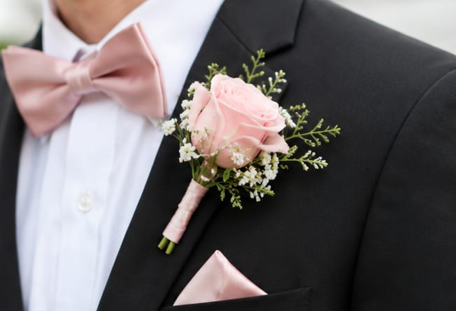 Simple Light Pink Boutonniere  - A pale pink rose boutonniere that reads romantic and timeless-perfect as a wedding boutonniere or for prom, anniversary, or any formal celebration. The soft pastel rose sits center stage, accented by small white filler blooms and fresh green sprigs so the person you're honoring looks polished and thoughtfully styled against a dark suit.  Neatly wrapped and secured with a boutonniere wrap, this single boutonniere is made for comfortable all-evening wear and easy pinning to lapels. The delicate mix of pink, white, and green keeps the look versatile for bridesmaids, groomsmen, fathers, or the guest of honor; it's shown paired with a pink satin bow tie and pocket square to illustrate a coordinated look, but this listing is for the boutonniere only. Order ahead to match party colors and request multiples for consistent, elegant presentation.