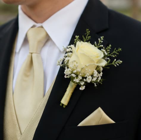 Simple Pale Yellow Rose Boutonniere  - A cream rose boutonniere that reads timeless and refined-perfect for a wedding boutonniere or other dressy celebration. The Simple Pale Yellow Rose Boutonniere pairs a single soft ivory-yellow rose with delicate white filler blossoms and fresh green sprigs to create an elegant accent that complements a suit jacket, pale gold tie, or pocket square. The understated palette of white, yellow, and green gives the person wearing it a quietly polished, classic look.  Carefully wrapped and wired for secure pinning, this boutonniere is sized to sit neatly on a lapel without overwhelming the jacket. Because it's made as a single-rose piece, it works beautifully for grooms, groomsmen, fathers, or anyone you want to feel especially seen on the day. Order as the boutonniere-only item; for best results, pin it to the left lapel and keep it cool and mist lightly until wear to preserve freshness.