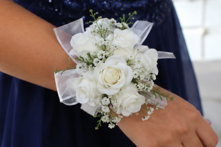 Simple White Corsage  - Clustered white spray roses and delicate filler blooms form a compact, rounded wrist corsage designed for weddings and other dressy occasions. As a wedding corsage, prom corsage, or formal accessory, its all-white palette and translucent ribbon loops create a timeless, elegant look that lets the person you're celebrating feel polished and perfectly styled.  The arrangement pairs small white roses with tiny complementary blooms and soft greenery for gentle texture and visual depth. Sheer ribbon adds a light, flowing accent while a comfortable wrist corsage band keeps the piece secure and wearable throughout the event. Sized to sit neatly on the wrist, this corsage is meant to complement bridal parties, attendants, or honorees without overpowering their outfit.  Give this corsage to the person you're thinking of when you want them to feel cherished and put-together for a ceremony, reception, or prom night. Because it's crafted for wear, order with the event time in mind so the flowers look fresh on arrival; if you need specific sizing for the wrist band, please include that in your order notes.