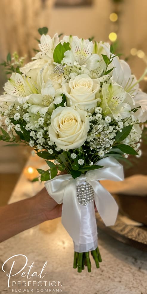 White Mixed Flower Nosegay - Creamy white roses take center stage in this white mixed flower nosegay, a compact hand-tied bouquet perfect for a wedding bouquet or romantic anniversary gift. The bridal-ready look combines white alstroemeria and delicate sprays of baby's breath for a soft, layered texture, while fresh green foliage peeks through to add natural contrast - the kind of bouquet that makes the person you're thinking of feel cherished and seen.  Tightly rounded and easy to carry, stems are wrapped in white satin ribbon and finished with a sparkling rhinestone brooch for an elegant, formal touch. Colors are primarily white and green with small mixed-color accents from the filler blooms; size is designed as a traditional nosegay for hand-held occasions. This arrangement arrives ready to tuck into a vase; keep stems trimmed and in fresh water to extend vase life for several days.