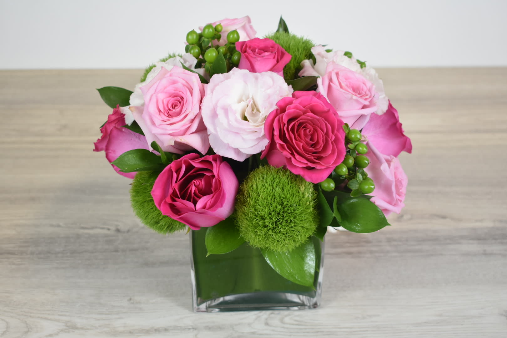 Compact arrangement of pink roses, white lisianthus, and green dianthus in a clear glass cube vase