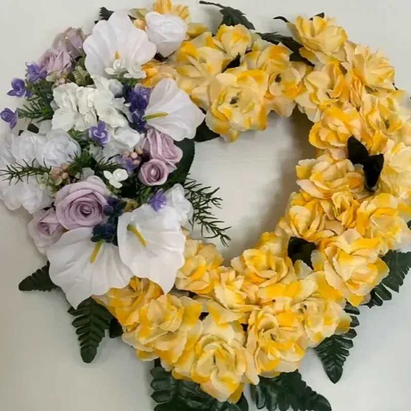 Memorial Wreaths X12 18" - A heart-shaped sympathy wreath that reads as a gentle, personal tribute-perfect for funerals or memorial services where you want to honor a loved one with warmth and grace. This funeral flower arrangement places yellow roses around the outer edge for a bright, comforting halo, while a mixed cluster on the left-white orchid-like blooms, lavender roses and small purple accents-adds a softer, more contemplative note that speaks to remembrance and love.  Handmade on an 18" heart-shaped wreath form, the design uses dense clusters of roses and orchids with dark green artificial foliage peeking through to define the silhouette and give long-lasting structure. The palette of yellow, white, purple and pink creates contrast and depth without feeling busy; textures range from the velvety rose petals to the smooth orchid shapes and delicate filler blooms. Because it's mounted on a sturdy form with artificial greenery, the wreath arrives ready to display and keeps its shape throughout services and viewings.  Choose this wreath when you want the person you're honoring to feel remembered and cherished-the bright yellow conveys warmth and celebration of life, while the lavender and white bring quiet comfort. The 18" size makes it suitable for chapel displays, graveside stands, or home memorials; minimal care is needed since the form and foliage maintain the arrangement's appearance during the memorial period.