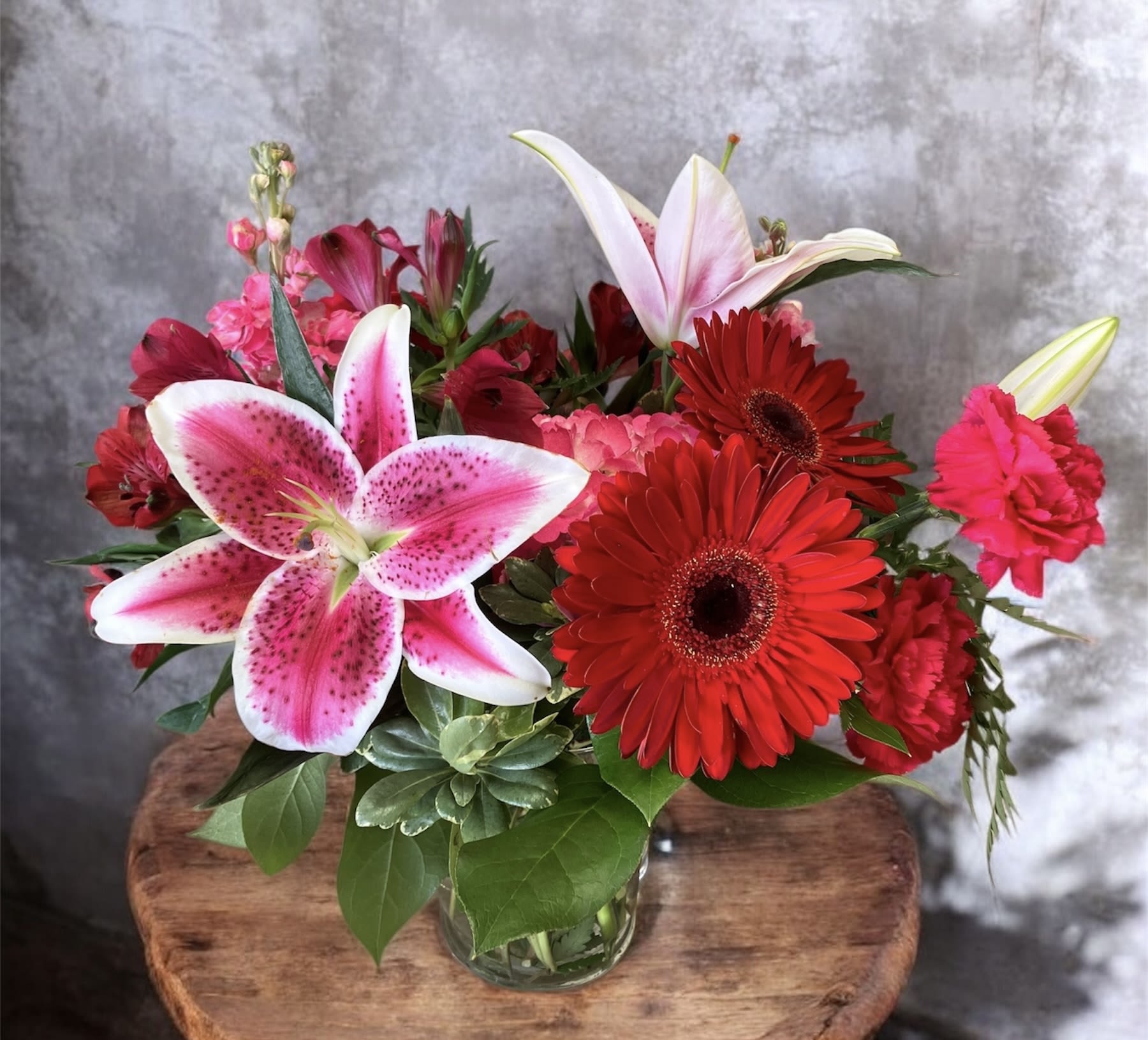Passion - A romantic, lush arrangement designed to captivate the heart. Vibrant red gerbera daisies create a passionate focal point, paired beautifully with hot pink carnations and stock for rich pops of color. Pink hydrangea adds fullness and balance, while graceful stargazer lilies lend elegance and a touch of intoxicating fragrance. Accented with Peruvian lilies, this arrangement feels affectionate and beautifully expressive, perfect for celebrating love.