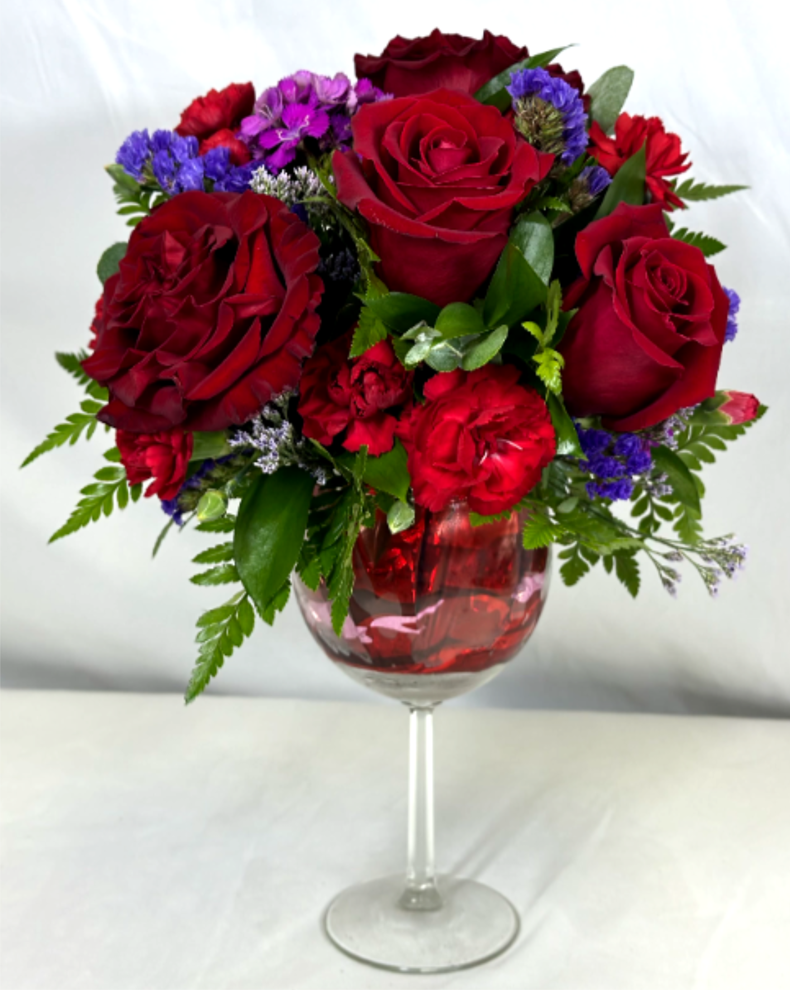 Raise a Glass. Red Wine - What better way to celebrate than with flowers and wine!? We will take care of the flowers with this beautiful arrangement in bright and deep red, designed in a large sturdy reusable wine glass. You supply the wine, and it will bring a smile for all. If the featured flowers are unavailable we will create your gift using the freshest available blooms of equal or greater value.