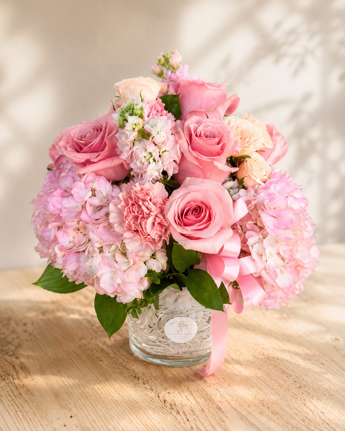Eirene's Tender Bloom – Luxury Pink Vase Arrangement - Inspired by Eirene, the Greek goddess of peace and gentle harmony, this arrangement reflects quiet elegance through soft pink blooms and graceful garden textures.  Designed in Flower Fairy's signature garden style, this luxury vase arrangement features lush pink hydrangeas, premium pink roses, delicate carnations, stock, spray roses, and carefully selected seasonal flowers. Layers of blush and soft pink blooms create a full, rounded design with timeless elegance, while the clear glass vase and satin ribbon complete its refined presentation.  Perfect for birthdays, anniversaries, Mother's Day, baby showers, thank-you gifts, congratulations, get well wishes, or simply reminding someone they are loved.  Each arrangement is handcrafted by Flower Fairy using the freshest flowers available. While occasional substitutions may be necessary due to seasonal availability, we always preserve the overall style, color palette, and premium quality of every design.  Features: Luxury vase arrangement Pink hydrangeas Premium roses Carnations Seasonal flowers Garden-style floral design Glass vase Handcrafted by Flower Fairy Same-day flower delivery in Phoenix