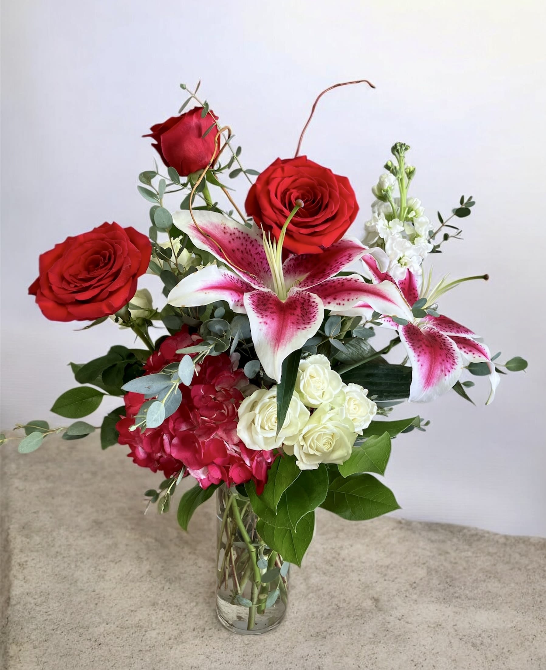 Sweetheart - A romantic floral arrangement showcasing stargazer lilies as the focal point, surrounded by rich red roses and lush red hydrangea for depth. White spray roses and white stock add softness and balance, while eucalyptus brings a fresh, natural accent. Flowing curly willow completes the design with graceful movement and an organic, elegant finish.