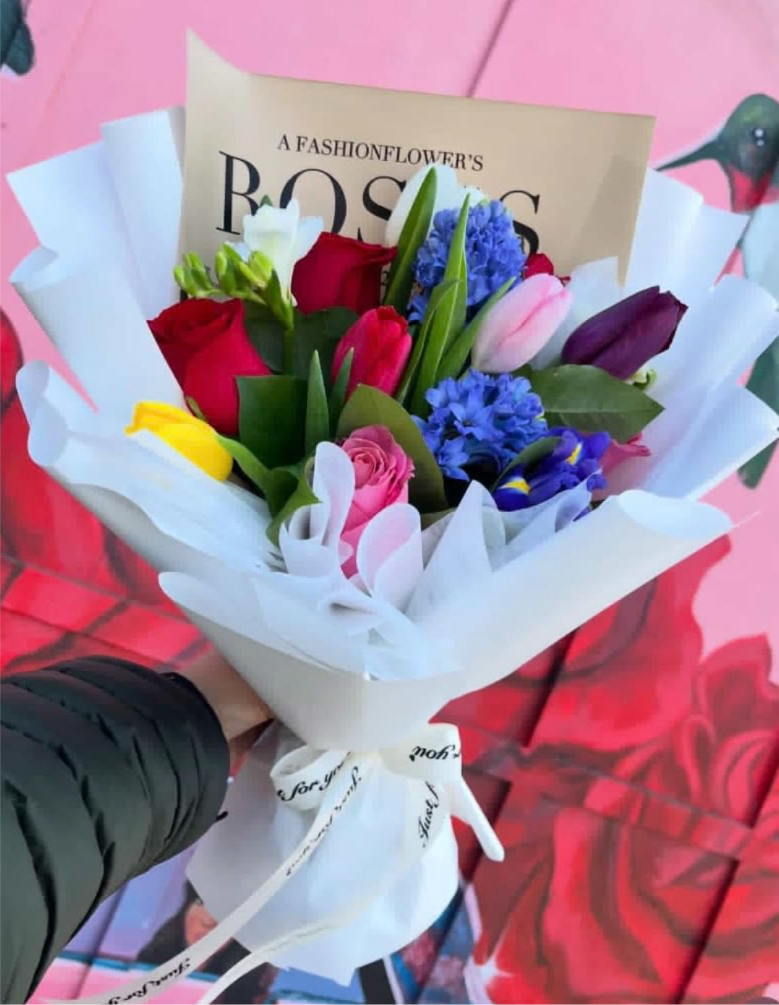 Rose Tulip and Hyacinth Korean Bouquet - A lush mix of red and pink roses, vibrant tulips, and clustered blue hyacinth-style blooms creates an immediate impression of joy and romance - ideal as a birthday bouquet, anniversary gift, or a bright "thinking of you" surprise. The saturated palette of red, pink, white, yellow, purple and blue reads celebratory and affectionate, so the person you're sending it to will feel genuinely noticed and celebrated.  Hand-tied in a compact, layered style, the arrangement features roses, tulips, hyacinth accents, freesia and iris for varied texture and scent. The stems are finished in a Korean-inspired white paper wrap and tied with a white ribbon; a printed card is included to add your message. The polished, gift-ready presentation makes it easy to order for personal celebrations, romantic moments, or congratulations - choose the size option that fits the occasion and enjoy same-day pickup or delivery where available.