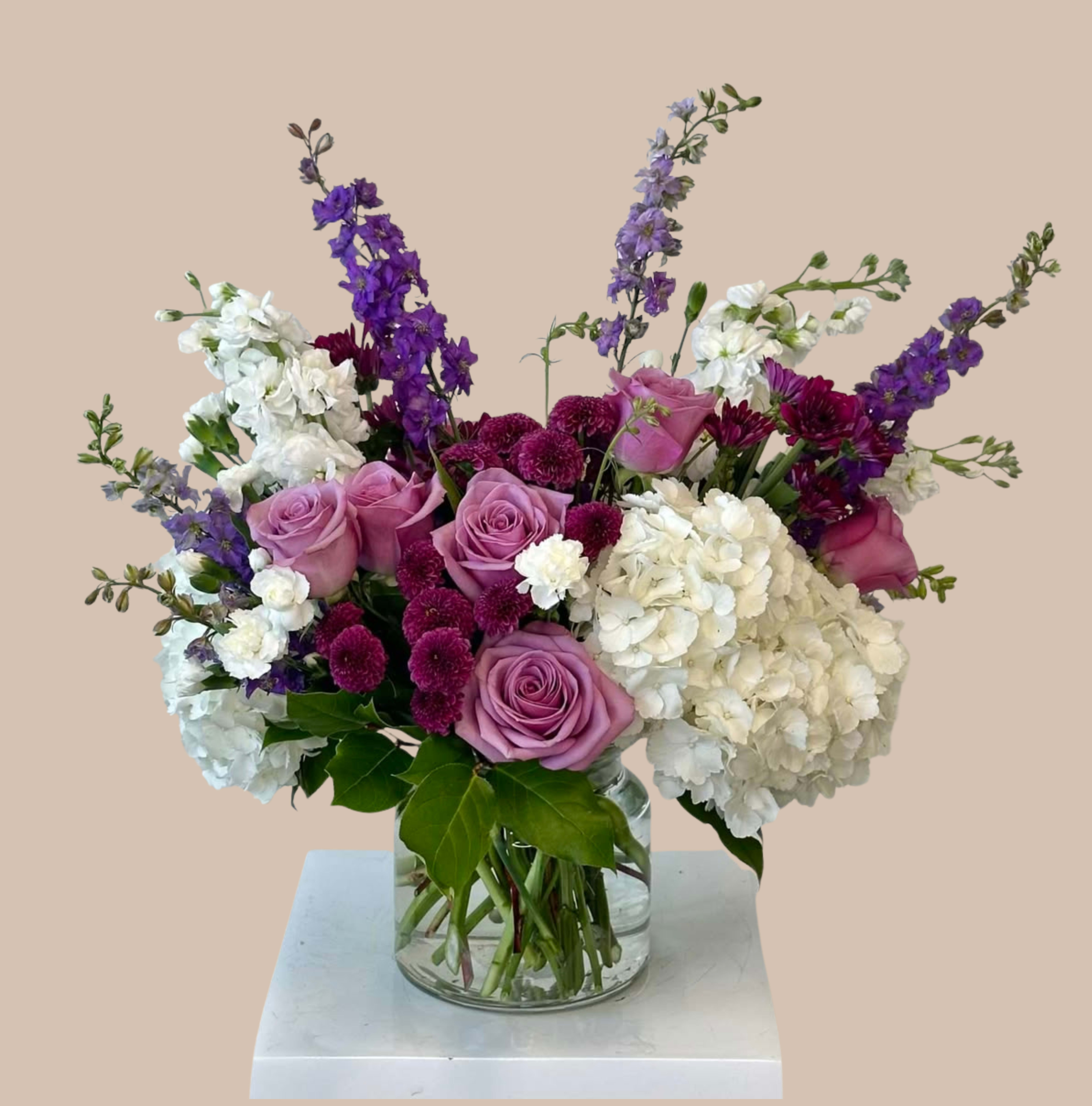 Stem &amp; Shadow Deluxe - White, lavender, and dark purple florals form the heart of this garden-style flower arrangement, a romantic anniversary bouquet that also reads beautifully for birthdays, thank-yous, or "thinking of you" moments. Tall spires of purple stock and delphinium rise above a rounded base of deep magenta chrysanthemums and soft daisies, giving the design an airy, layered silhouette that feels both elegant and generous-the kind of arrangement that makes the person you're sending it to feel truly seen.  Creamy whites, lilac and lavender tones, vivid purples and touches of pink and green play across the composition for a balanced, textured look. Arranged in a clear glass vase, the mix of roses, hydrangeas, stock, delphinium, chrysanthemums and daisies offers contrast between soft, full blooms and vertical accents for lasting visual interest. Sized to sit on a dining table or bedside, it's equally appropriate for "I'm sorry," get well, sympathy, or love and romance gestures; provide fresh water and trim stems occasionally to keep the display looking its best.