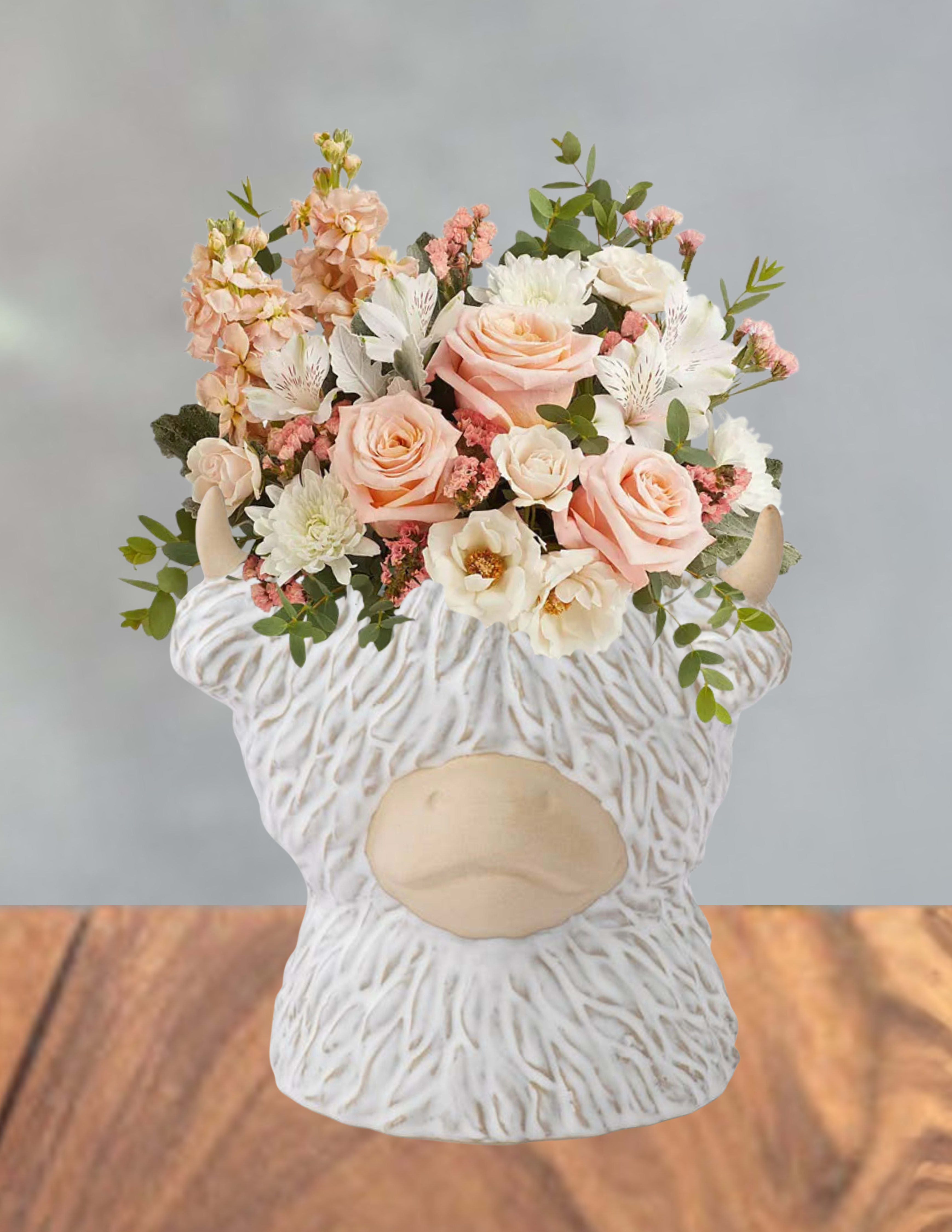 Peaches and pastures - Soft peach roses and blush blooms gathered in a compact, rounded bouquet give this flower arrangement a gentle, romantic feel-perfect as a birthday bouquet, a springtime gift, or a cheerful "thinking of you" surprise. The design pairs white alstroemeria and pale chrysanthemums with small pink filler flowers to create soft layers of cream, pink, and green that read tender and uplifting for anniversaries or to brighten a feminine home.  Flowers are arranged tightly for a lush, tactile look and arrive nestled in a white textured ceramic vase shaped like a whimsical bull head, adding playful character to the classic color story. The ceramic vessel doubles as a keepsake accent for mantel or entryway, while the mix of roses, alstroemeria, chrysanthemums and delicate fillers provides varied texture and soft fragrance.  For the person you're celebrating, this arrangement sends a message of warmth and lighthearted affection-she'll feel noticed and cherished. The compact size makes it easy to display; refresh water daily and recut stems every few days to extend bloom life. Order soon for spring delivery or to brighten someone's day any time of year.