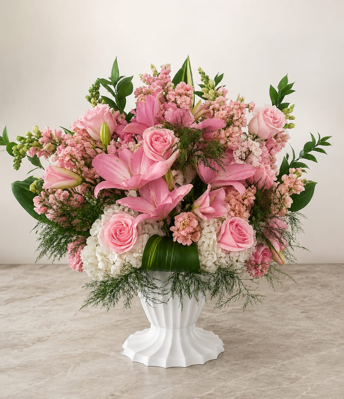 Garden of Tenderness - Soft pink roses and lilies take center stage in this Garden of Tenderness flower arrangement, perfect as a birthday bouquet, anniversary flowers, or an elegant thank-you gift. The pastel pinks and crisp whites create a romantic, gentle impression that makes the recipient feel truly cherished the moment they see it.  A full, rounded design layers pink roses and pink lilies with clusters of pale pink stock and white hydrangeas for lush texture and scent. Soft greenery frames the composition as the blooms rise above a classic white pedestal vase, giving the arrangement a graceful, sculpted silhouette that sits beautifully on a dining table or entry console.  Made to feel both special and approachable, this arrangement suits celebrations and everyday gestures alike-she'll feel seen and celebrated whether it's a milestone or a simple surprise. The compact, symmetrical shape arrives ready to display in its white pedestal vase; refresh water as needed and trim stems slightly when rewatering to help blooms last.