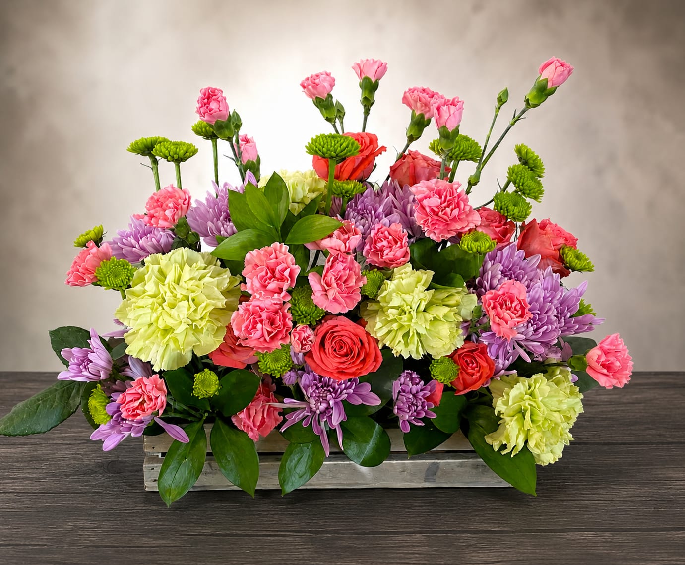 Field of Flowers - A lush, garden-style centerpiece overflowing with color, this flower arrangement is perfect as a birthday bouquet, spring celebration centerpiece, or just-because gift to brighten someone's day. The low, rectangular wooden box gives the piece a rustic, hand-tended feel while the compact, layered design makes an immediate cheerful impression-the person you're thinking of will feel truly celebrated by its abundant, joyful energy.  Packed tightly for a full, rounded silhouette, the design mixes pink carnations, purple chrysanthemums, red roses, and pale green and soft yellow carnations (or chrysanthemums), all nestled among glossy greenery. The bright pinks and violets pop against chartreuse and pale yellow accents, creating lively contrast and garden-inspired texture. Finished in a rectangular wooden box, this arrangement sits low and wide-ideal for tabletops, mantels, or as a festive centerpiece.  Perfect for spring occasions, birthdays, anniversaries, or an everyday surprise, it sends a message of warmth and cheer. The compact, dense styling keeps blossoms supported and looking fresh; standard care like fresh water and a cool spot will help prolong vase life.