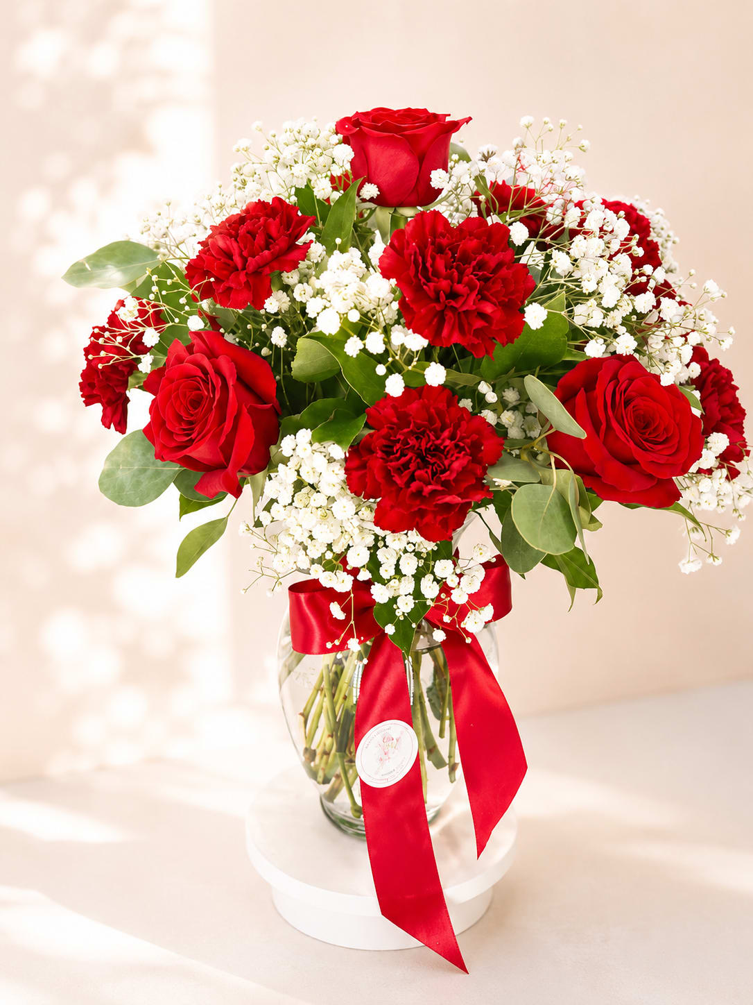 Carmenta's Crimson Promise – Vase Arrangement - Inspired by Carmenta, the Roman goddess of cherished beginnings and heartfelt blessings, Carmenta's Crimson Promise celebrates enduring love with a classic combination of velvety red roses, lush carnations, airy baby's breath, and fresh greenery. Designed in a clear glass vase and finished with Flower Fairy's signature ribbon, this arrangement is both elegant and unforgettable.  Each arrangement is handcrafted using the freshest flowers available, ensuring every design is naturally unique while maintaining its timeless romantic style.  Perfect for anniversaries, Valentine's Day, birthdays, romantic surprises, or any occasion when words aren't enough.  Features: Premium red roses Fresh red carnations Baby's breath Seasonal greenery Clear glass vase Handcrafted by Flower Fairy Same-day flower delivery throughout the Phoenix Metro Area Designed by your local Phoenix florist