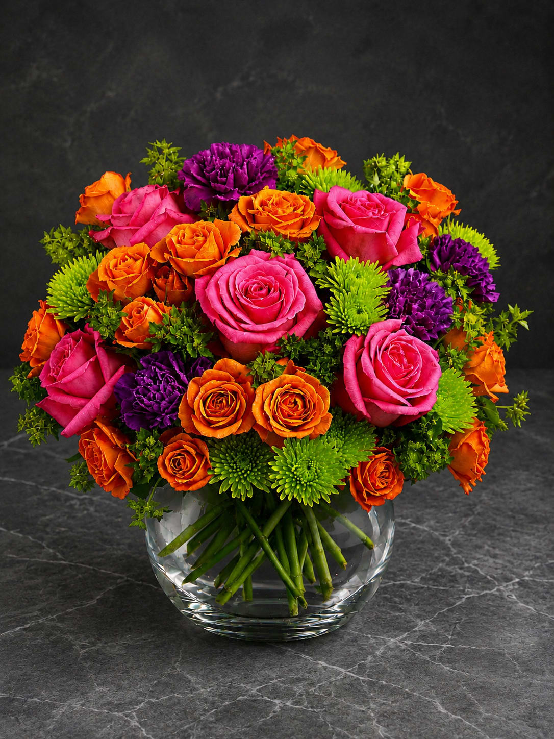 Tropical Radiance - Hot pink and orange roses take center stage in this vibrant birthday bouquet, balanced by vivid purple blooms and bright green chrysanthemum pompons for a cheerful, celebratory look. The rounded, densely packed shape reads energetic and bold-the kind of arrangement that makes the person you're thinking of feel truly seen and celebrated on their special day.  Arranged in a clear glass vase that keeps the presentation clean and modern, the stems are visible below for a fresh, natural feel. The mix of roses and chrysanthemums brings a pleasing contrast of velvety petals and textured pompons; colors include hot pink, tangerine orange, jewel-toned purple, and lime green for maximum impact. Ideal as a birthday gift, congratulations bouquet, or just-because surprise, this arrangement arrives ready to display and brighten any room.