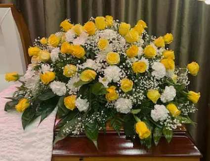 Eternal Garden - Bright yellow roses set against a cushion of white carnations make a luminous, respectful statement in this casket spray designed for sympathy and funeral services. The Eternal Garden arrangement brings a hopeful, uplifting touch-perfect for honoring a life with warmth while remaining appropriately formal for a memorial setting.  This low, full spray lays across the casket with a rounded silhouette: clusters of bright yellow roses scattered through a base of pure white carnations and delicate white filler flowers, all framed by glossy dark green foliage for depth and contrast. The palette of yellow, white, and green reads both vibrant and serene, and the composition is crafted to present beautifully from front pews or in photographs.  Offer this as a way to express sympathy, celebrate joyful memories, or bring light to a solemn service-the person you're honoring will feel genuinely remembered. For sizing and placement details or to coordinate additional pieces, please contact us so we can ensure the spray fits the casket and the tone of the service.