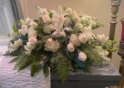 Peaceful in White - A soft, layered sympathy arrangement designed to bring calm and comfort, Peaceful in White is a large, low floral design ideal for memorial services or to send as a thoughtful sympathy gesture. This floral arrangement and sympathy centerpiece mixes roses, lilies, carnations, hydrangea clusters, and delicate filler blooms in white, pink, cream and touches of green and blue, creating a traditional, serene presence that helps the person you're thinking of feel seen and supported.  Arranged in a low rectangular container, the composition spreads gracefully with broad fern fronds and glossy foliage for fullness and gentle texture; the overall shape reads formal and restful, suited to a service table or placement beside a casket. Expect a generous size and layered, garden-like look-soft petals, subtle fragrance from the lilies, and a calm palette that reads beautifully from a distance while offering comforting detail up close.  For sympathy and remembrance, this is the kind of arrangement that quietly honors a life and offers solace to family and friends. If you need delivery timed to a funeral or service, please let us know the date and location so we can ensure timely placement.