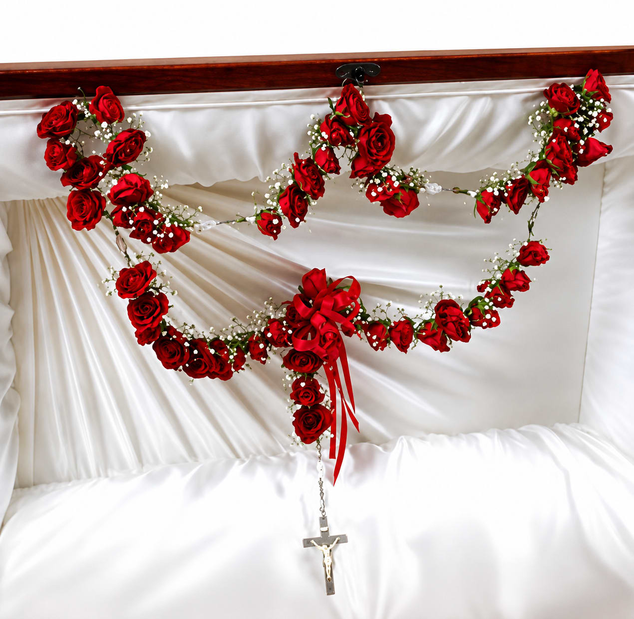 Rosary Arangememt  - A draped garland of roses and delicate white filler flowers laid across a satin fabric creates a solemn, ceremonial tribute perfect for a funeral or memorial service. The design includes a gathered ribbon bow at the lower center with a small crucifix suspended beneath, giving the piece a reverent, comforting presence that honors the person you're remembering.  Made with mixed roses and complementary blooms, the arrangement rests on a white satin drape and is finished with a tasteful ribbon and crucifix. The sweeping, formal composition is crafted to be seen from the front, with textures that combine the lush rose heads, airy filler flowers, and smooth satin for a composed, dignified effect.  This arrangement is intended to convey sympathy and respect at a funeral or memorial; it's the kind of tribute that helps the bereaved feel supported and remembered. Please note any placement or delivery instructions when ordering so we can ensure it arrives ready for the service.