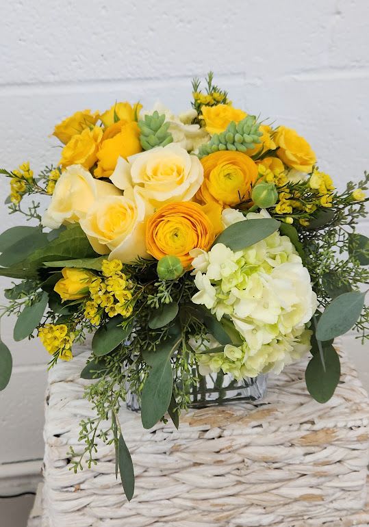 Low yellow and cream flower arrangement with roses, ranunculus, hydrangea, and succulents in a glass cube vase