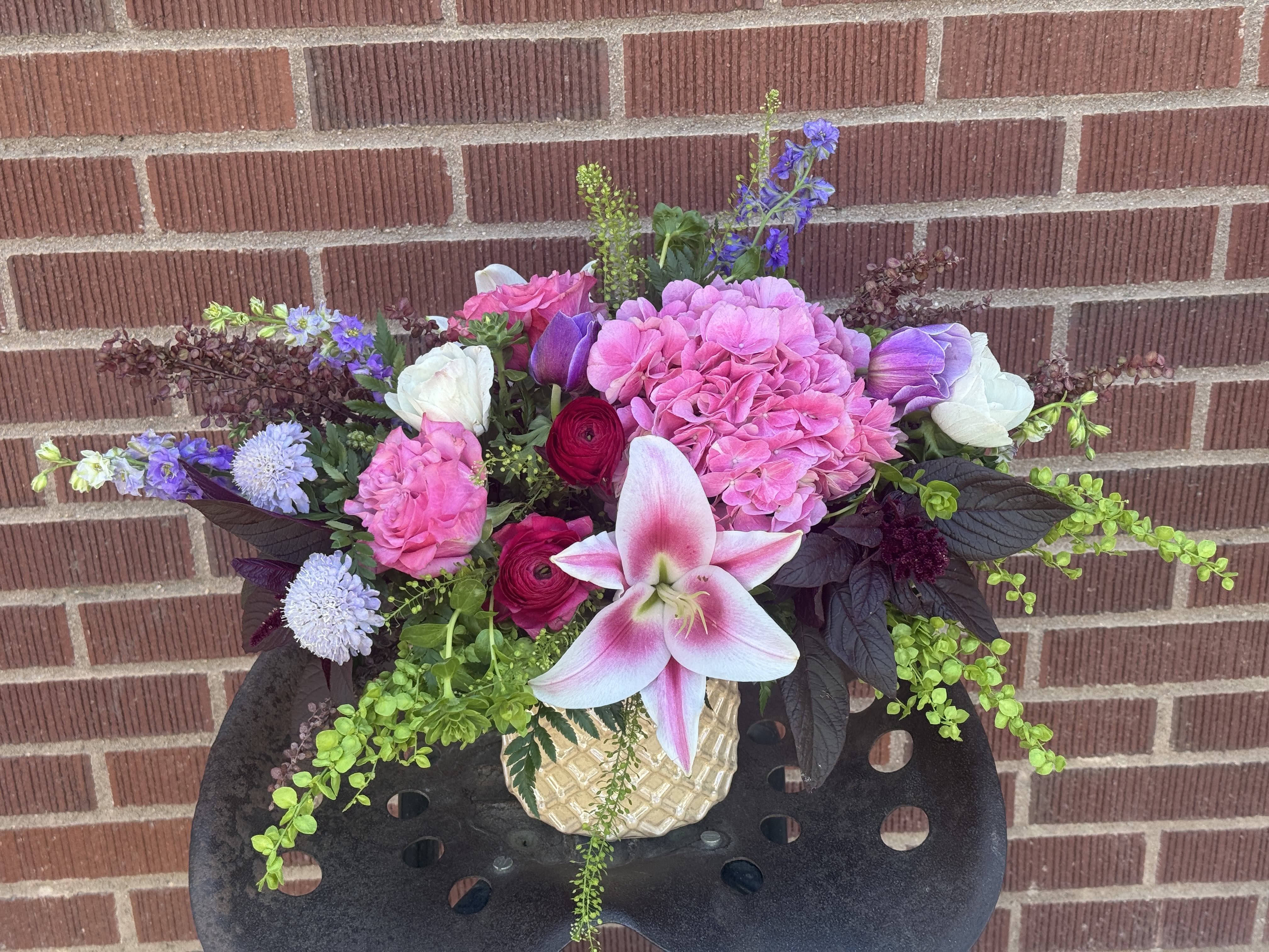 Bohemian Rhapsody - LIMITED EDITION, WEEKLY DROP! - This striking, custom-designed masterpiece showcases an intentionally dense and wild gathering of premium seasonal stems. A magnificent starburst oriental lily serves as the focal crown, offset by a pillowy dome of antique pink hydrangea. Sculptural columns of cobalt delphinium and trailing chartreuse tendrils break traditional boundaries, introducing dramatic height and movement. Deep crimson ranunculus and ruffled magenta garden roses add dense layers of saturated color, nestled securely in a textured, woven-pattern stone jar. It offers a contemporary, asymmetrical aesthetic designed to captivate from every angle.