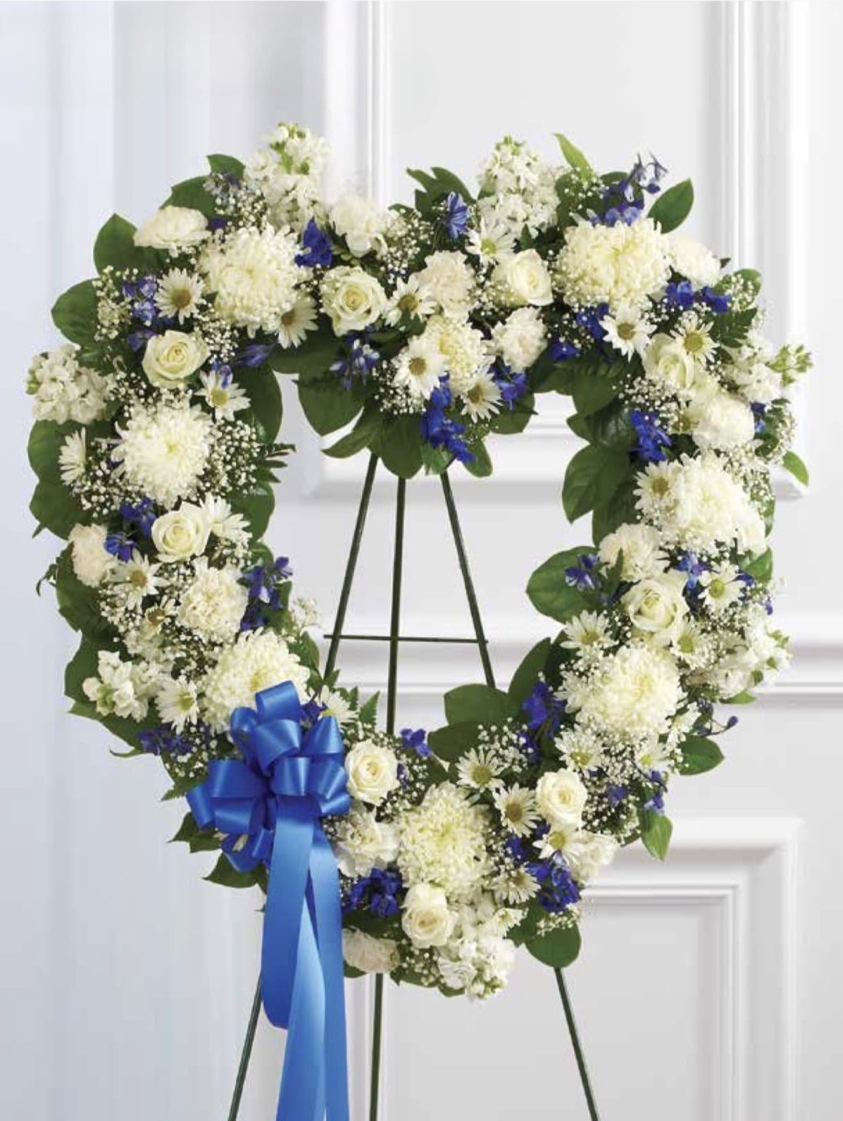 White with Blue Open Heartq - A large heart-shaped funeral wreath arranged on a metal easel, crafted in white and vivid blue for a respectful memorial or romantic tribute. The white with blue open heart is designed to convey sympathy or lasting love-the heart silhouette and ceremonial blue ribbon bow make it especially fitting for a funeral service, graveside remembrance, or a meaningful anniversary tribute where solemn beauty matters.  Dense layers of white roses, white daisies, chrysanthemums and complementary blooms are set against broad green foliage, with striking blue accents woven throughout to create high-contrast visual impact. The arrangement reads formal and substantial from a distance; the metal easel provides secure display and the blue ribbon bow at the lower left adds a thoughtful finishing touch.  Made to stand tall at services or to grace a graveside, this heart wreath offers a dignified, heartfelt message for the person you're honoring. No special care is needed beyond standard service placement; order with delivery to the service location or funeral home for timely presentation.