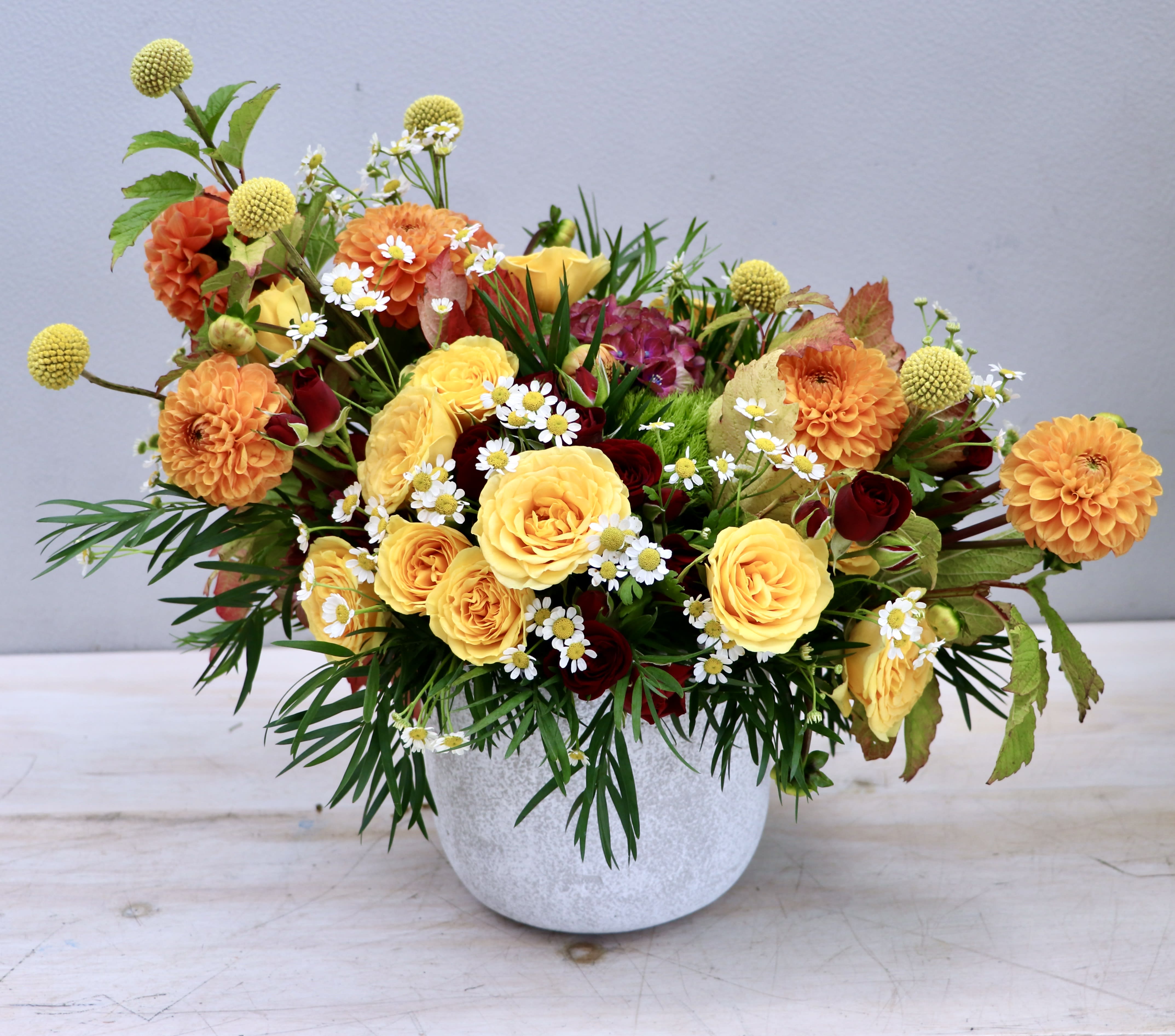 Low arrangement of yellow roses, orange dahlias, and small white daisies in a white pot