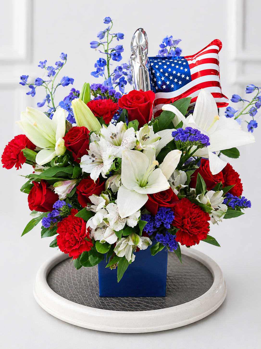 Honor remembrance tribute  - A striking patriotic floral arrangement featuring premium red roses, elegant white lilies, white alstroemeria, vibrant blue delphinium, carnations, statice, and lush greenery, accented with a patriotic balloon. Designed in a modern blue container, this arrangement is a beautiful way to honor military service members, veterans, first responders, or celebrate patriotic holidays such as Independence Day, Memorial Day, and Veterans Day.