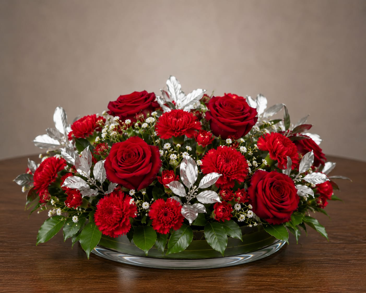 Silver Passion - A tightly clustered low event arrangement of vivid red roses and red carnations creates an immediate impression of elegance and celebration-perfect as a wedding centerpiece, anniversary table accent, or formal event floral. The compact, rounded profile sits in a wide glass bowl so the person you're honoring will feel the moment elevated without blocking conversation or sightlines.  Creamy white filler blooms and green accents thread through the red mass for fresh contrast, while metallic silver foliage gives the design a polished, festive edge. The composition rises just above the bowl rim for a full look; materials include roses, carnations, assorted greenery, and decorative silver leaves arranged densely for symmetry and visual impact.  Designed to sit low on reception tables or a special mantel, this arrangement is the kind of floral that makes someone feel celebrated and seen. Final size matches a standard wide glass bowl presentation-ideal for multiple place settings or a focal table-and requires simple care: fresh water in the bowl and a cool location away from direct sun to keep the blooms looking their best through your event.