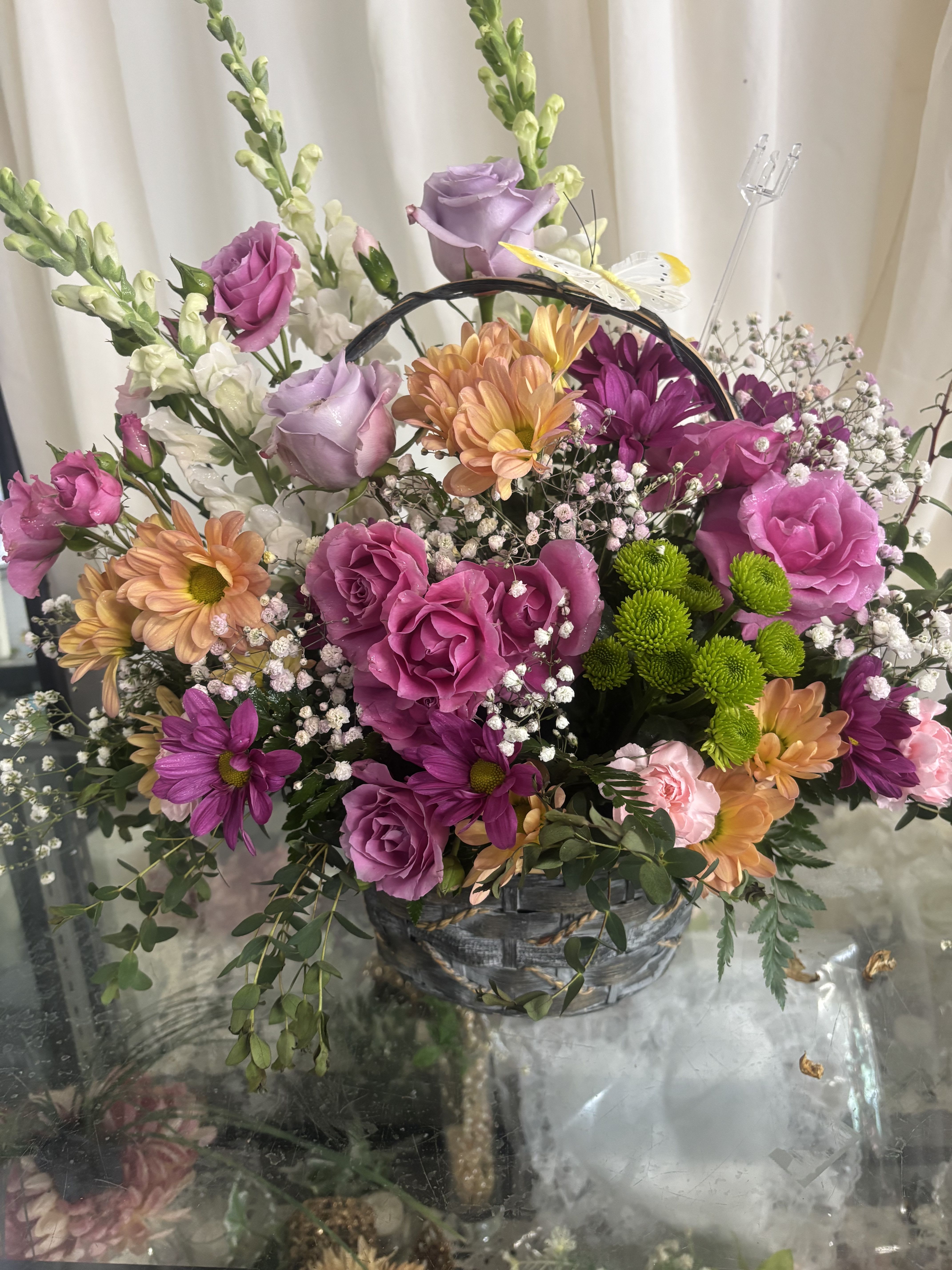 Blooming Basket Case  - This full design pairs roses, carnations, lilies, daisies, chrysanthemums, snapdragons and delicate white filler (baby's breath) for abundant texture: large round blooms anchor the composition, chrysanthemum-style flowers add volume, and tall snapdragon spikes give height and movement. 