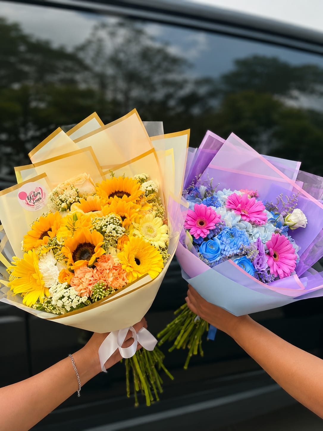Golden Sunshine Bouquet or Blue Harmony Bouquet - Bright, hand-tied bouquets that feel like a little celebration in someone's hands-the Golden Sunshine bouquet and the Blue Harmony bouquet are perfect birthday bouquets, congratulations flowers, or a just-because pick-me-up. The Golden Sunshine option centers on bright yellow sunflowers and gerbera daisies with soft peach and cream accents to create a warm, sunny impression; the Blue Harmony option pairs vivid pinks, cool blues, and lavender for a playful, uplifting contrast that still reads cheerful and refined. Either choice makes the recipient feel seen and celebrated the moment they open it.  Each arrangement is full and rounded, hand-wrapped in layered translucent paper and finished with a decorative ribbon for an elegant, ready-to-gift presentation. Flowers include sunflowers, gerbera daisies, roses, carnations, and complementary seasonal stems to add texture and depth in mixed color palettes of yellow, pink, purple, and blue. These bouquets are designed to arrive fresh and wearable as a hand-held gift; to keep them looking their best, place stems in water as soon as possible and recut stems at an angle before arranging in a vase. Order now for a bright, instant mood-lifter that suits birthdays, congratulations, or simply saying "I'm thinking of you."