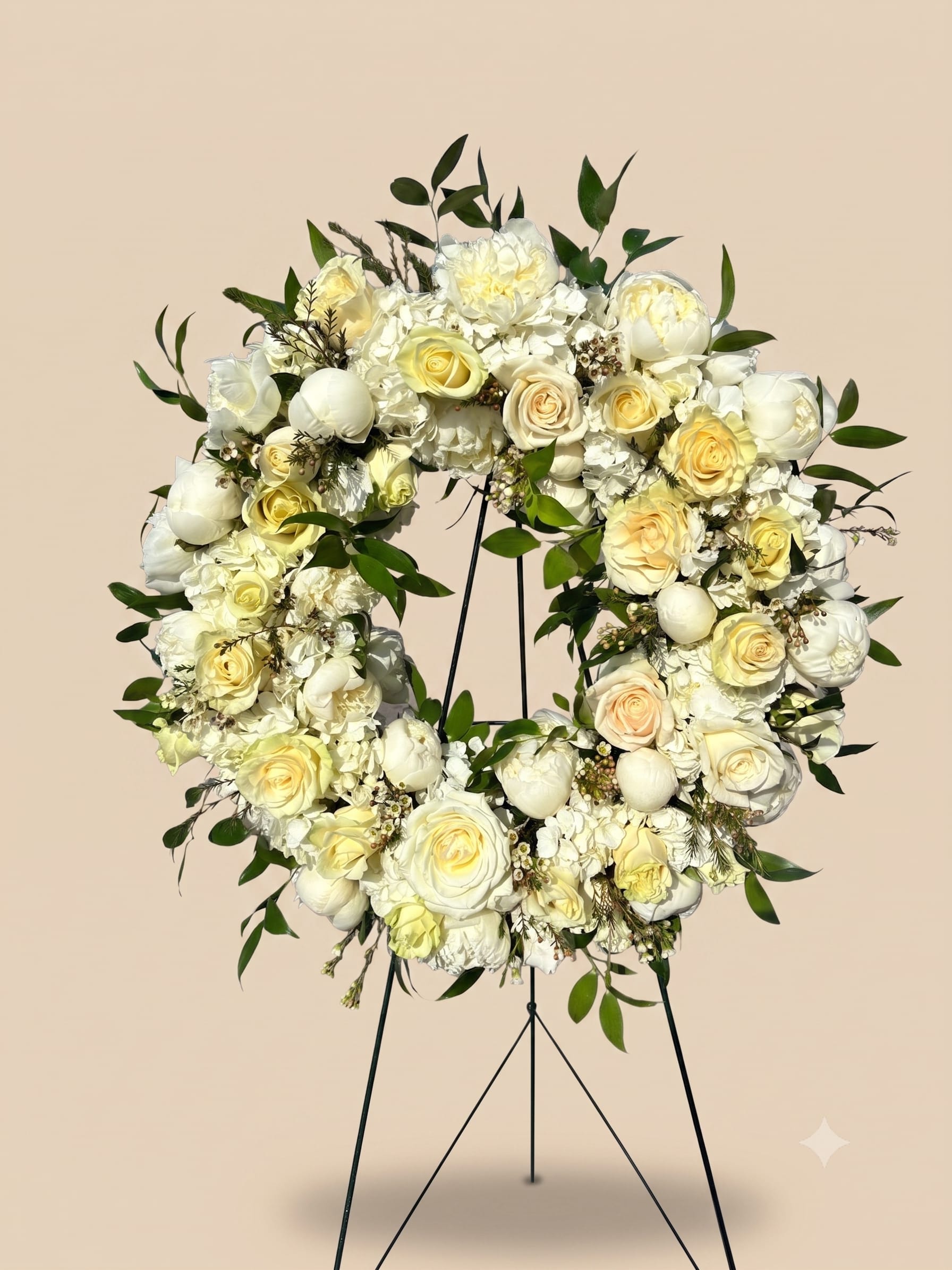 BP131 - Tranquil Ivory Bloom - A textured standing wreath featuring of creamy whites and soft pale yellows. This elegant, garden-style tribute is packed with premium blooms like lush roses, delicate peonies, and hydrangeas, beautifully accented with dynamic seasonal greenery.   Seasonality: The arrangement shown features Peonies (available late spring to mid summer).