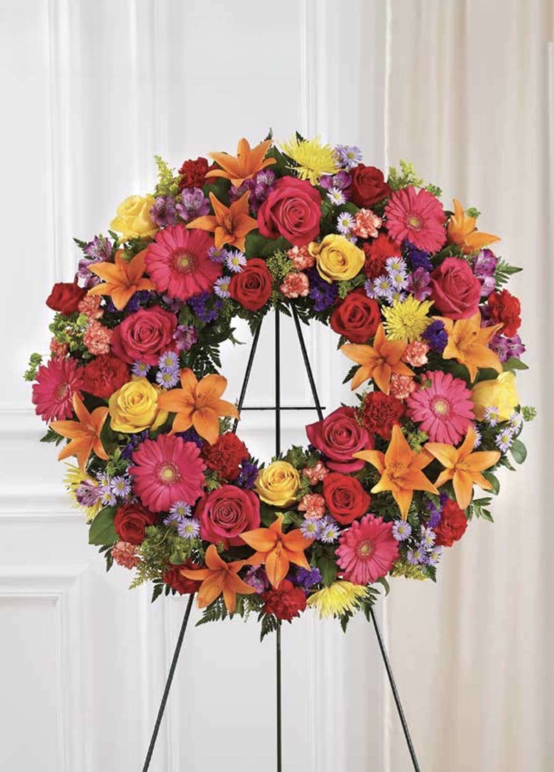 Bright Colorful Wreath - A bold wreath of bright pink roses and gerbera daisies paired with orange lilies, yellow roses, red carnations, and purple aster accents makes a vivid sympathy arrangement that celebrates a life with warmth and color. This flower arrangement for memorials and tributes hangs on a metal easel stand, delivering an uplifting, high-contrast statement that reads respectful and hopeful rather than somber-perfect when you want the person you're honoring to be remembered with joy.  The circular design is dense and symmetrical, crafted for strong visual impact from every angle. Expect a layered mix of roses, lilies, gerbera daisies, chrysanthemums, asters, and carnations in pink, orange, red, yellow, and small purple filler blooms, mounted securely on the easel for display. The scale and fullness are intended for formal services or graveside presentations where a substantial, colorful wreath is appropriate.  Choose this bright, colorful wreath when you want to send a message of celebration and remembrance-ideal for funerals, memorial services, or to place at a graveside. The florist prepares it ready-to-display on the included black metal easel; for delivery timing or special requests (color balance, personalization, or inscription ribbon), please contact the shop when ordering.