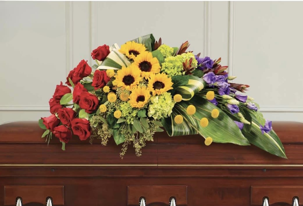 Colorful Casket Spray - A bold, full-top casket spray that brings warmth and color to a funeral or memorial service, this flower arrangement balances reverence with a celebratory spirit. The large, low floral spray is designed to rest across the top of a wooden casket, making it an ideal sympathy tribute that honors a life with vivid reds and sunny yellows alongside more contemplative purples and greens. The person you're remembering will be seen and celebrated-this is the kind of arrangement that says your memories are bright and cherished.  Carefully layered for visual depth, the spray features classic red roses paired with bright yellow sunflowers, purple blooms, clustered green accents, and broad tropical leaves for a lush base. The composition reads formal and ceremonial without feeling purely somber-the contrasting colors create striking visual interest while the low profile keeps the arrangement dignified and viewable from all sides. Sized to span the length of a standard casket, it presents a full, even silhouette that complements traditional services and outdoor celebrations alike.
