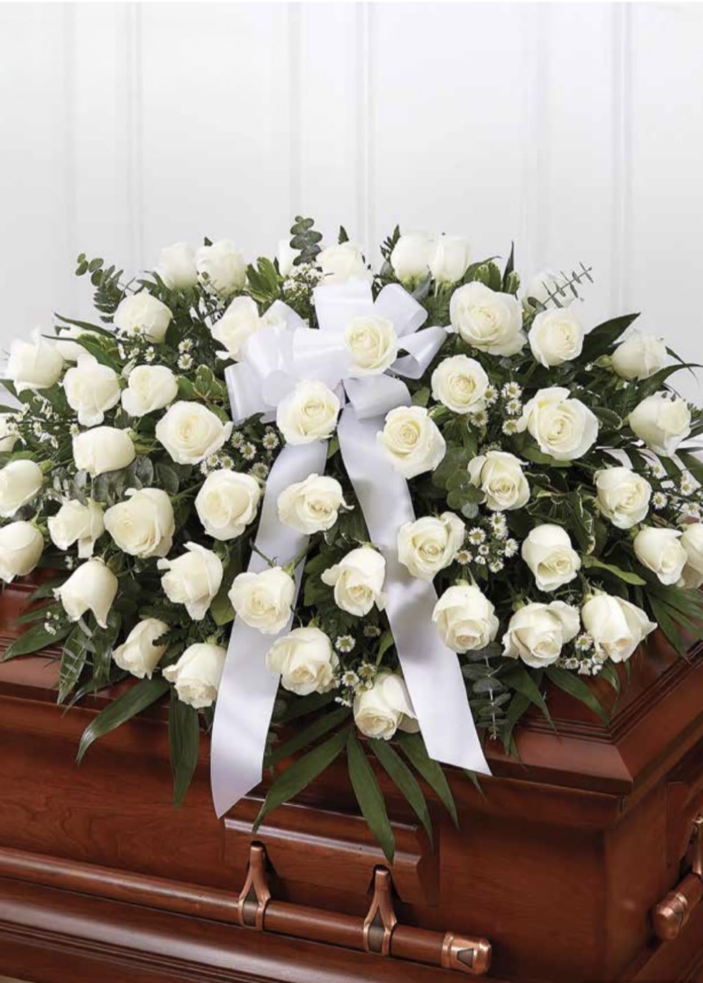 White Rose Casket Spray - A serene cascade of densely clustered white roses creates the centerpiece of this white rose casket spray, designed specifically for funeral and sympathy services. The low, spreading shape sits across a polished wooden casket for a traditional memorial presentation, and the central wide white ribbon lends a formal, restrained touch that honors the gravity of the moment. This arrangement is meant to make the person you're thinking of feel tenderly remembered and deeply respected.  Composed mainly of pure white roses with small filler flowers and mixed greenery, the spray combines soft texture and crisp contrast-velvety petals against airy greens-to convey calm and consolation. Sized to drape naturally over a standard casket, it offers full coverage without overwhelming the setting. The white ribbon provides a dignified focal point, and the overall color palette of white and green keeps the presentation timeless and appropriate for a range of services.