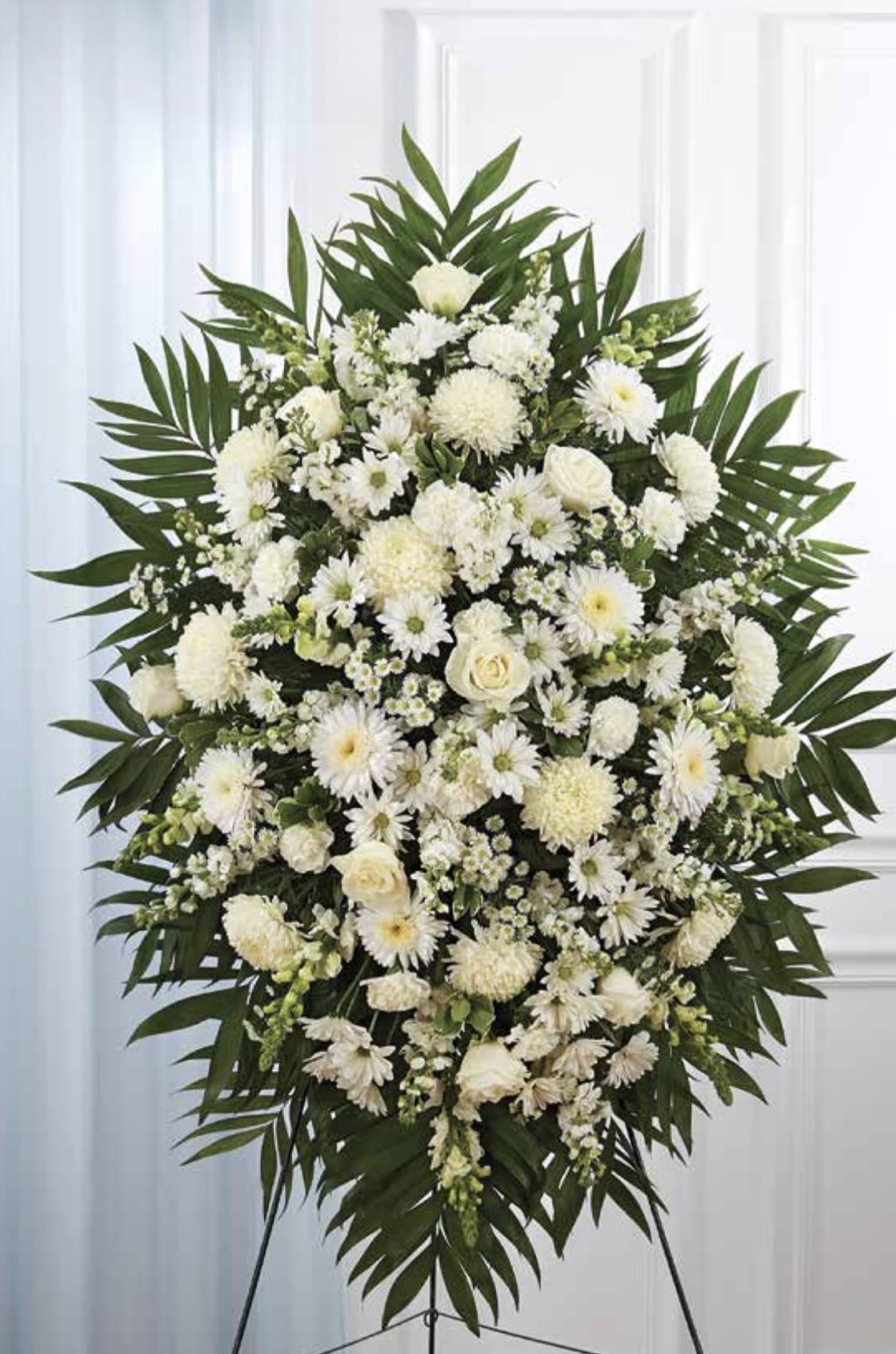 White Standing Spray - A serene all-white standing spray crafted for sympathy and funeral remembrance, designed to offer a graceful, respectful tribute. This white standing spray reads with formal dignity-perfect for honoring a loved one or for display at a memorial service-its calming palette and structured silhouette help convey sympathy and quiet support to the person you're remembering.  The arrangement features a dense mix of white roses, daisy-like blooms, and rounded chrysanthemums, arranged on a floral stand and framed by broad palm fronds that form a strong triangular shape. Lush green foliage provides contrast and vertical height, while the combination of round and layered textures gives the spray visual depth from every angle. Sized for prominent placement at a service, it presents a substantial presence without relying on color.  Made to comfort and to mark a meaningful farewell, this spray is suited for a funeral home or graveside display and sends a clear message of reverence and sympathy to the bereaved. If you need help selecting delivery timing or placement instructions for the service, please let us know so we can ensure it arrives appropriately.