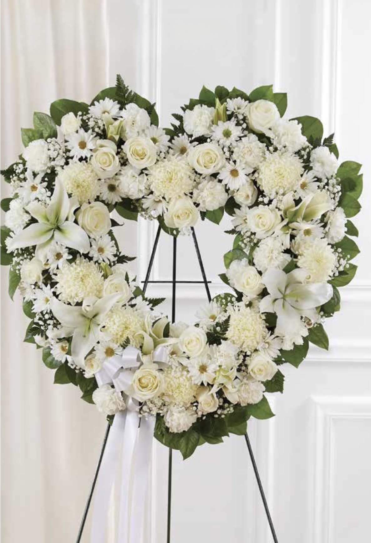 White with Lilies Open Heart - A large, heart-shaped sympathy arrangement in serene whites-this open heart wreath makes a respectful statement for a funeral or memorial service. The white lilies and roses at the center draw the eye, while the soft daisy-like chrysanthemums and carnations add gentle texture; it's the kind of tribute that will help the person you're thinking of feel honored and remembered.  This floral arrangement uses a layered mix of white roses, lilies, daisies, carnations and clustered white filler blooms set against deep green foliage that frames the heart shape. Mounted on a metal easel stand and finished with a simple white ribbon bow, the piece reads formal and understated. The design is full and substantial; it's clear silhouette and monochrome palette keep attention on the sentiment rather than the show.  Designed specifically for sympathy and memorial moments, this wreath conveys deep sympathy and quiet respect for family, friends, or a beloved colleague. Because it arrives on an easel, it's ready for service display; no additional care is required beyond keeping it in a cool place until the event.