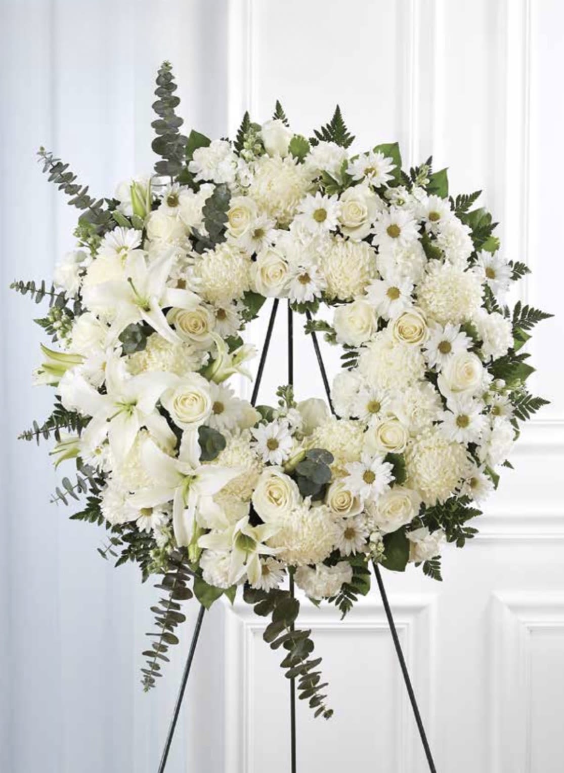 White with Lilies Wreath - A serene circular sympathy wreath in white and green, centered on graceful lilies and classic white roses, offers a quiet, dignified tribute for a memorial or funeral. This white floral arrangement for sympathy or remembrance creates an immediate impression of calm - the kind of display that helps the person you're honoring feel respectfully remembered.  The wreath is densely packed with white roses, lilies, daisies, and full chrysanthemum blooms set against eucalyptus and rich green foliage for contrast and texture. Its large, symmetrical form reads formal and traditional; the dark green leaves frame the white flowers so their shapes and soft petals stand out. The wreath is presented on a sturdy metal easel stand for easy display at a service or graveside.  Choose this arrangement when you want the recipient's family to feel comforted and seen - a respectful way to express sympathy and support. The easel-mounted design is ready to place at a memorial service; minimal care is needed once it's arranged on site, and local delivery can ensure timely placement for services.