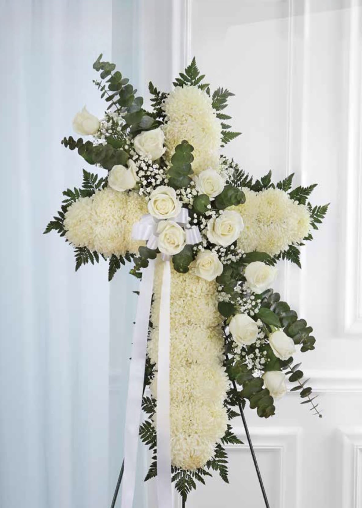 White Cross with White Rose Cluster - A large white standing cross designed for sympathy and memorial services, this floral arrangement communicates reverence and comfort with quiet elegance. The funeral cross arrangement features dense white chrysanthemum or carnation blooms forming the cross, accented across the face with a cluster of white roses and sprigs of baby's breath - a traditional, dignified choice for honoring a loved one.  Dark green foliage frames the edges to give definition and contrast against the all-white palette, while white ribbon streamers cascade from the center for a ceremonial finish. Arranged on a floral stand and sized to serve as a prominent easel tribute, it offers a soft, understated presence that helps the person you're thinking of feel remembered and honored.