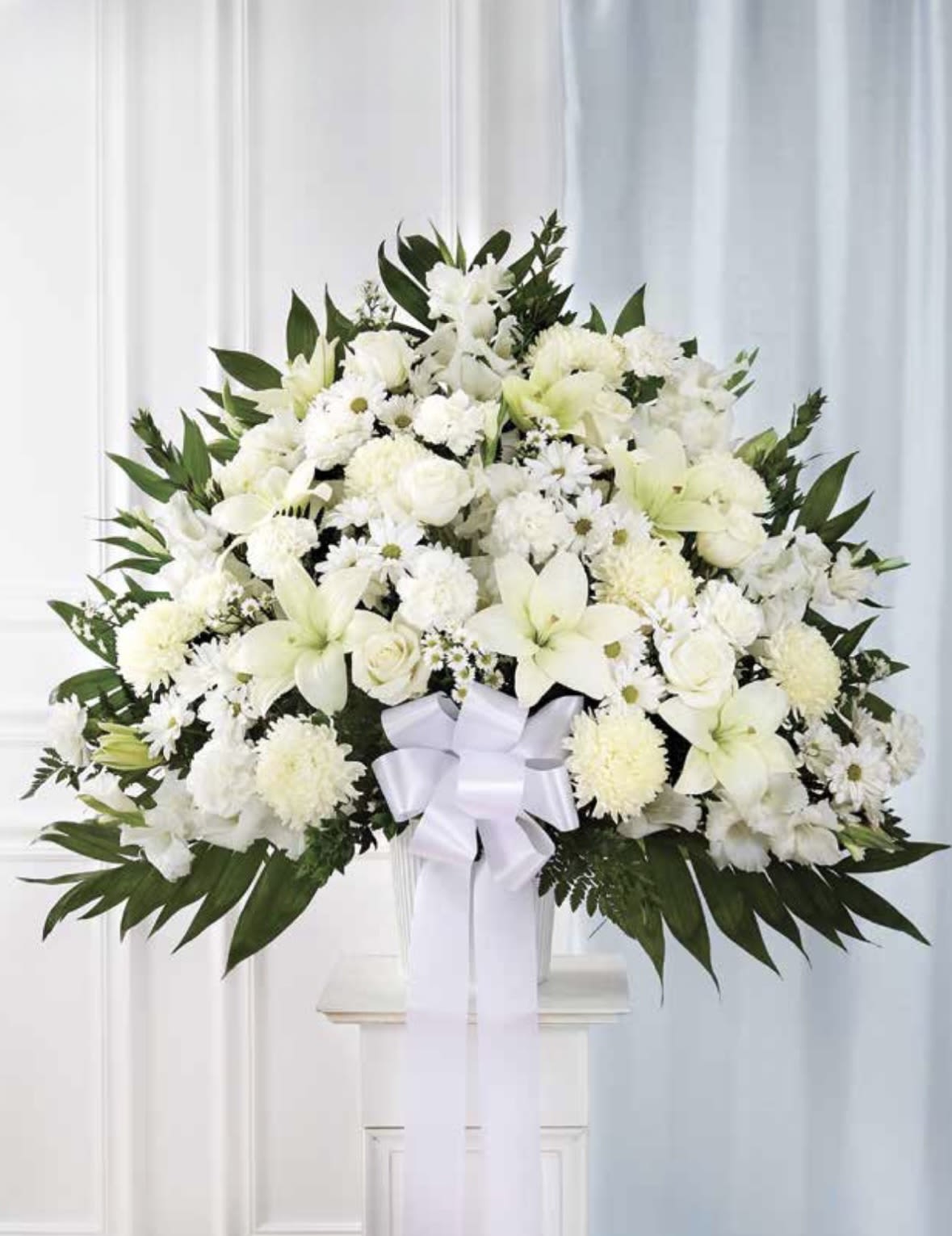 White with Lilies Pedestal Arrangement - A serene, formal pedestal arrangement crafted for sympathy or funeral service. This white pedestal arrangement centers on pure Asiatic lilies and ivory roses to convey quiet reverence. The arrangement reads as a classic white funeral arrangement, the kind that lets the person you're honoring feel truly remembered while offering comfort to those who gather.  Dense clusters of white chrysanthemums and daisy-like blooms are layered with the lilies and roses for texture and a rounded, substantial silhouette, while deep green foliage frames the composition for clean contrast. A wide white ribbon bow hangs at the center and the piece, making it suitable for chapel viewings or graveside memorials.  Designed to express sympathy with dignity, this arrangement is the thoughtful choice when you want the recipient's family to feel supported and seen. It arrives ready to display; for best presentation keep flowers in a cool, sheltered spot until the service and follow any local care instructions provided at delivery.