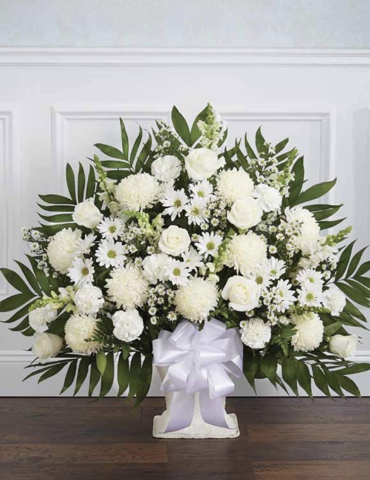 White Pedestal Arrangement - A serene white pedestal arrangement designed for sympathy or memorial services, this floral tribute offers a traditional, comforting presence that helps express your condolences. The formal white arrangement and pedestal vase create an understated, respectful centerpiece for a funeral service or graveside memorial, conveying sympathy and gentle remembrance to the person you're honoring.  Layered blooms of white roses, rounded chrysanthemums, daisy-like flowers and tall snapdragons form a full, textured surface framed by broad tropical-style greenery for a graceful, structured silhouette. The design sits on a classic pedestal vase and is finished with a white satin ribbon bow at the base, giving the piece a clean, elegant profile and substantial vertical height appropriate for viewing at services.  Ideal for comforting a grieving family or marking a solemn occasion, this arrangement reads as both peaceful and dignified-she'll feel remembered and honored when this is placed in the service space. Available in a large, symmetrical composition; please call the shop with any special requests or delivery details to ensure proper placement at the service.
