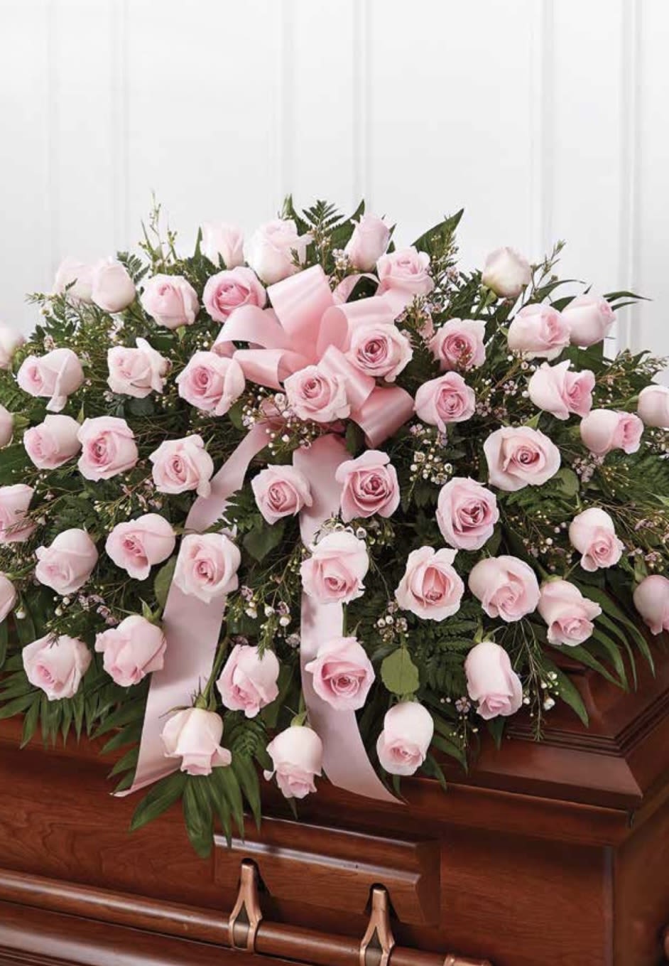 Pink Rose Casket Spray - A soft cascade of pale blush roses creates a comforting, traditional casket spray perfect for funeral or memorial services. This pink rose casket spray announces sympathy and reverence with a romantic pastel palette-pink roses layered over rich greenery and finished with soft pink satin ribbon-so the person you're honoring will be remembered with tenderness and grace.  The arrangement is low and spreading, designed to sit neatly atop a wooden casket; roses are densely clustered for a full, symmetrical presentation that reads as a formal service piece rather than an everyday bouquet. Colors are pink and green, with satin ribbon woven through the design to add a gentle, solemn accent and a polished finish.  Choose this spray when you want to express sympathy, love, or tribute in a timeless, respectful way-the arrangement is sized for casket top display and crafted to arrive ready to place. For ordering help or to confirm placement timing for a service, our team at Merced Floral can assist with delivery and care details.