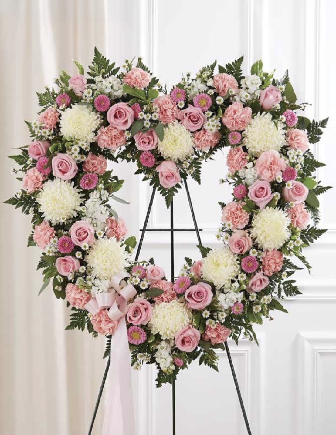 White and Pink Open Heart - A soft, open heart of pale pink roses and carnations framed by white chrysanthemums-this heart-shaped funeral wreath on a metal easel is a gentle, romantic sympathy arrangement designed to honor a loved one. As a memorial tribute or for a sentimental celebration of life, the pink and white palette reads of comfort and affection; the arrangement conveys that the person you're thinking of was cherished and will be remembered with tenderness.  The surface is full and layered, built from roses, carnations, chrysanthemums and small daisy-like blooms in shades of pink, white, and green for subtle depth. Displayed on a sturdy metal easel, the design includes a pale pink ribbon at the lower left for a quiet decorative accent. Its large heart shape makes a clear visual statement at a service or graveside while remaining tasteful and refined.  Choose this piece when you want the recipient's family to feel deeply seen and comforted-an unmistakable expression of love and remembrance. For delivery and placement at services, note the arrangement arrives ready on the easel; contact us with service details so we can ensure timely arrival and proper handling.