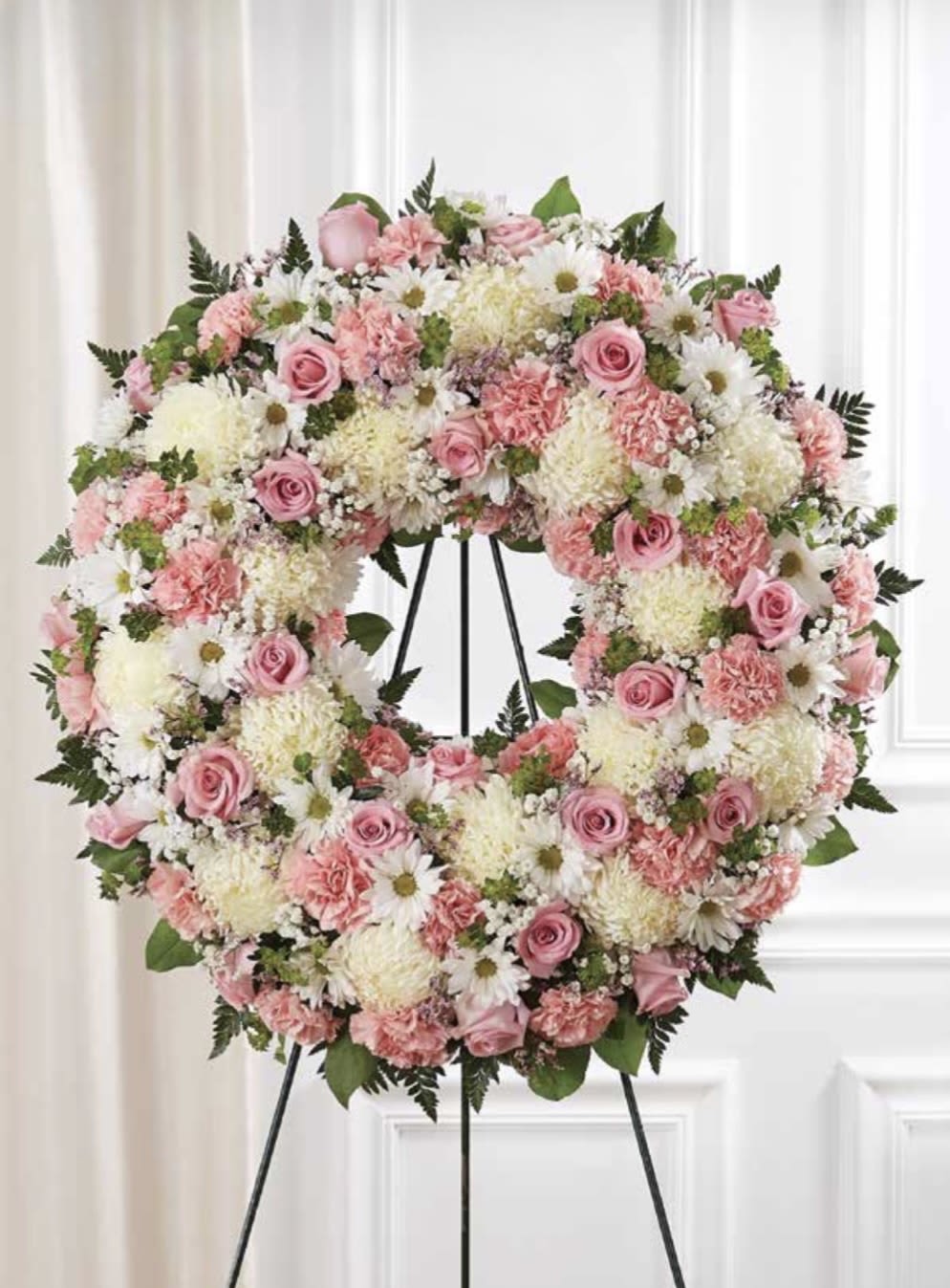 White and Pink Wreath - A soft, full circular wreath in pink, white, and green creates an immediate sense of gentle elegance - perfect as a sympathy arrangement or for a remembrance service. This white and pink wreath reads as both comforting and refined, the kind of floral tribute that helps the person you're honoring feel truly seen and remembered.  Closely packed pink roses and carnations form the wreath's romantic core, while pale chrysanthemums and white daisy-like blooms add delicate texture and light. The design is layered and plush, with green foliage peeking through to give subtle contrast and a natural finish. The arrangement is presented on a metal easel stand for a formal, dignified display that holds its shape and presence at services or memorials.  Appropriate for funerals, memorials, or any occasion calling for a tasteful floral tribute, this wreath sends a message of sympathy and care without overwhelming the moment. Order with the recipient and service details in mind - the easel stand is included for immediate display - and expect a composed, long-lasting arrangement that maintains its soft color palette throughout the service.